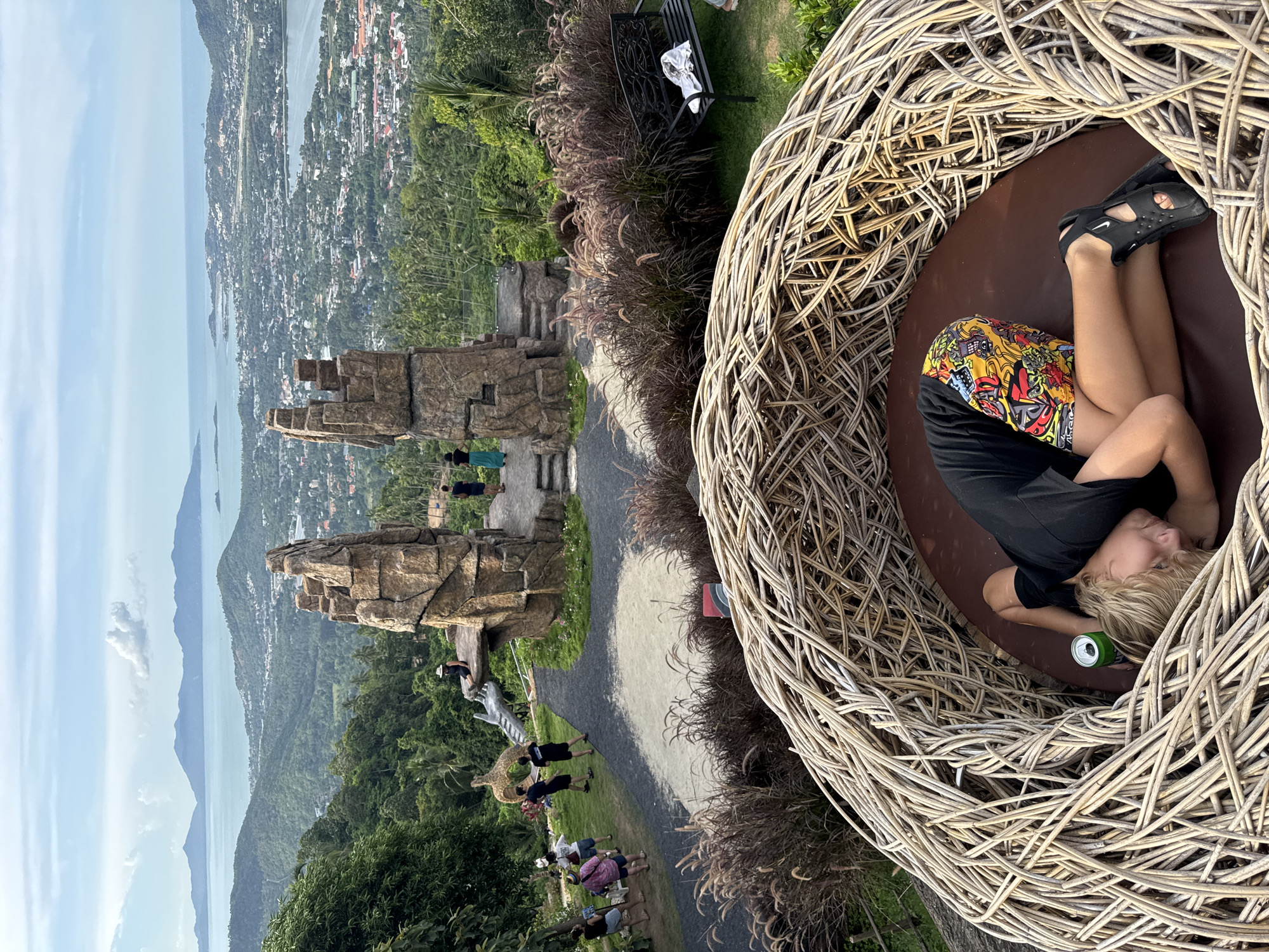 In the foreground, a blonde person lies on their side within a large, woven nest-like structure, their head resting on a brown cushion. They are wearing a black t-shirt and brightly colored shorts, with black sandals on their feet. A green can is visible next to their head. In the mid-ground, there are people standing around a rocky, architectural structure that resembles ancient ruins. Beyond that, the scene opens up to a panoramic view of a city nestled amongst lush green hills, with a body of water and distant islands under a cloudy sky. The environment appears to be an outdoor scenic viewpoint.