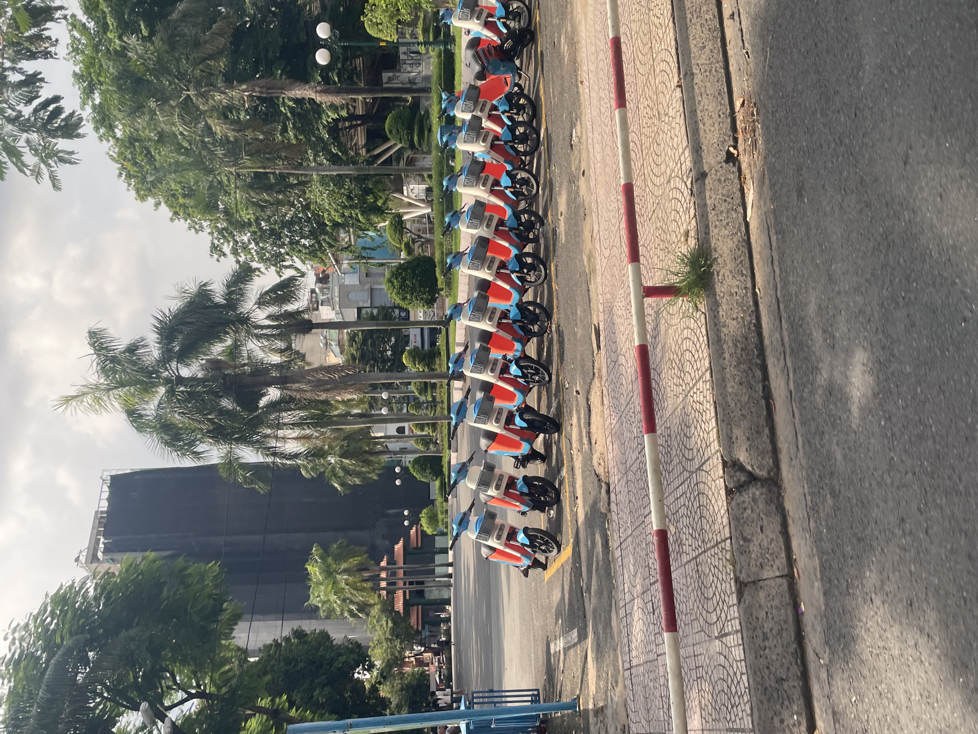 The image shows a row of electric scooters parked on a paved area in Thủ Đức, Vietnam. The scooters are predominantly white and orange with blue accents and are lined up neatly. In the background, a tall building under construction or renovation is visible, covered in dark sheeting, surrounded by palm trees and other lush greenery. Other buildings and street lights are also present in the distance. A red and white striped barrier separates the sidewalk from the street in the foreground. The overall scene depicts an urban environment with a focus on transportation and infrastructure development.