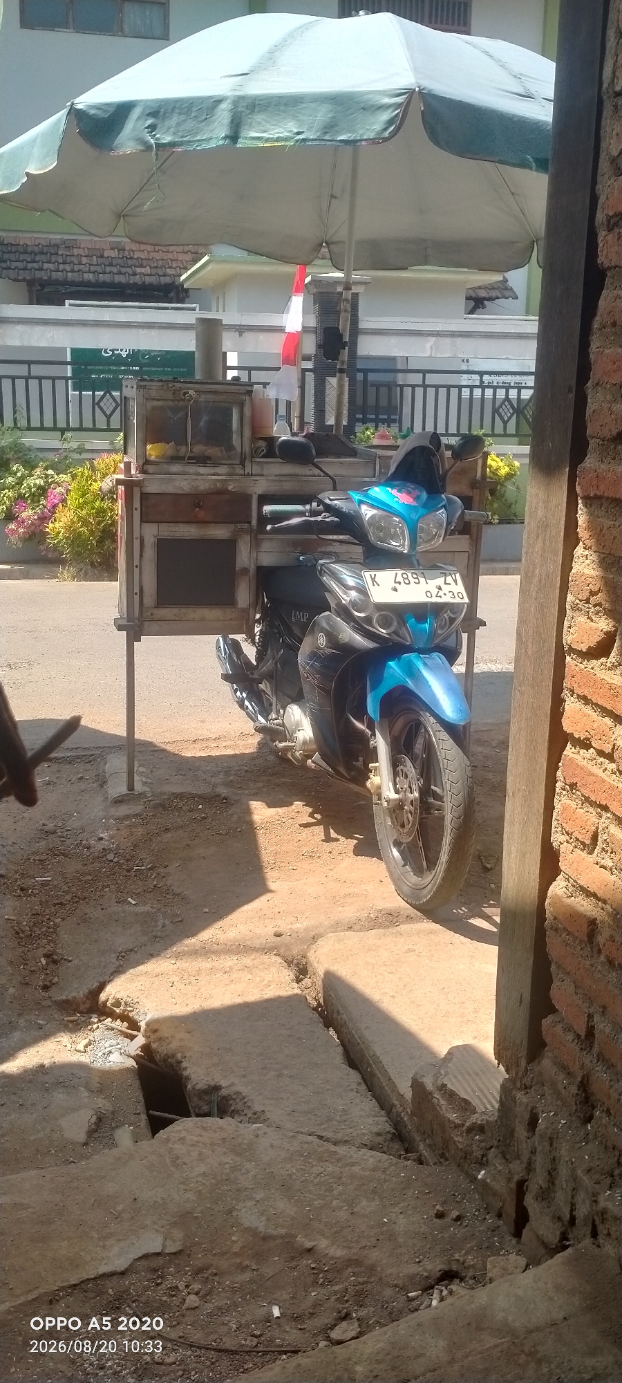 A blue Yamaha motorcycle is parked next to a wooden food cart under a large green umbrella. The food cart is supported by metal poles and contains various items within glass compartments. A license plate on the motorcycle reads "K 4891 ZV 04-30". The scene appears to be on a street, with a brick wall to the right and cracked concrete pavement in the foreground. In the background, there is a fence, some bushes with pink flowers, and buildings. The image includes a timestamp in the lower left corner: "OPPO A5 2020 2026/08/20 10:33". The overall environment suggests an outdoor setting in a city or town.