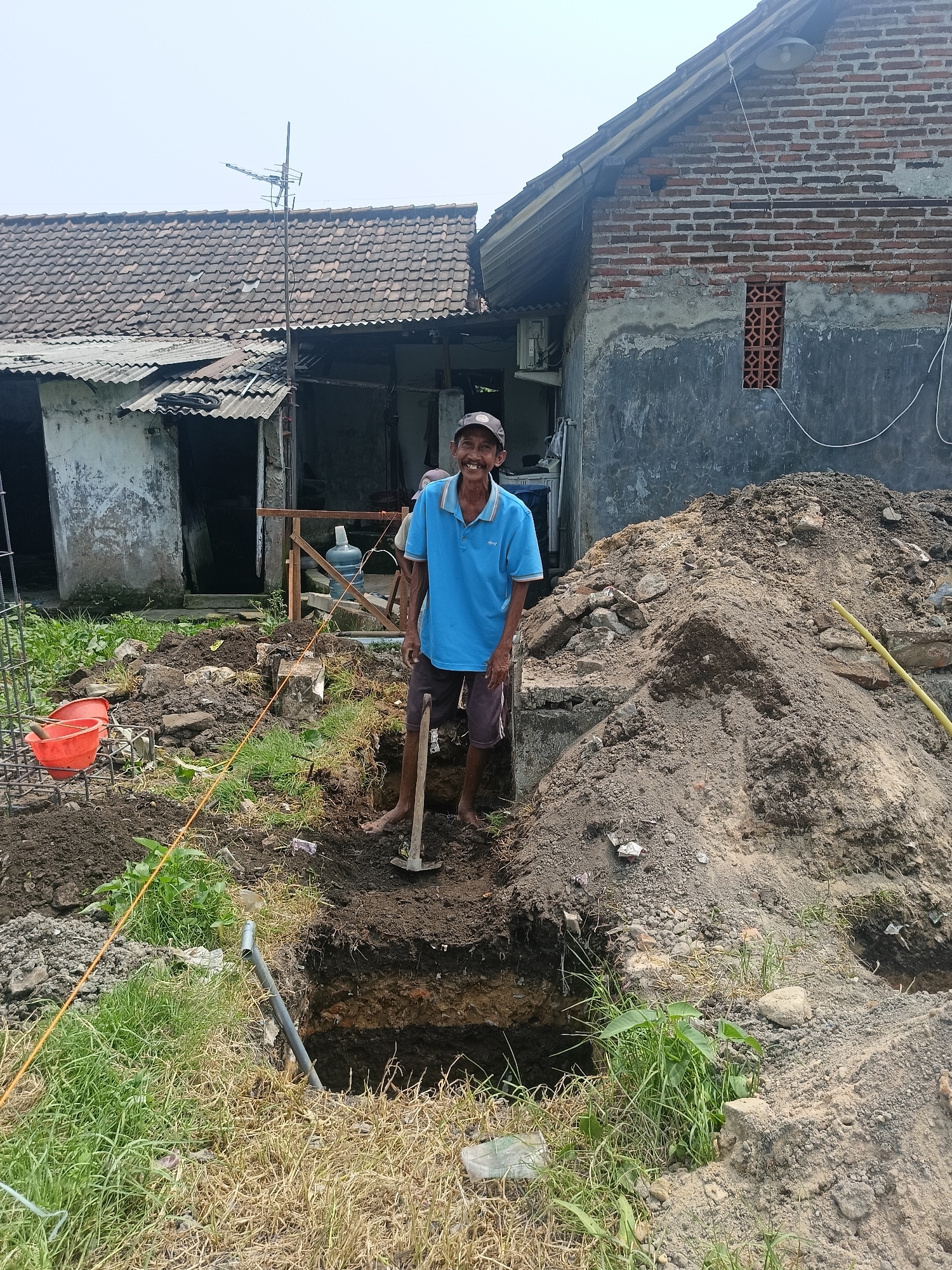 In Gedangan, Indonesia, a man stands by a construction site. He is wearing a blue polo shirt and dark shorts. He is holding a shovel and smiling at the camera. Behind him, a pile of dirt and rocks looms, with a partially dug hole in the foreground. The scene is set against the backdrop of two houses, one made of brick and the other of corrugated metal. The sky is overcast, suggesting a humid day.