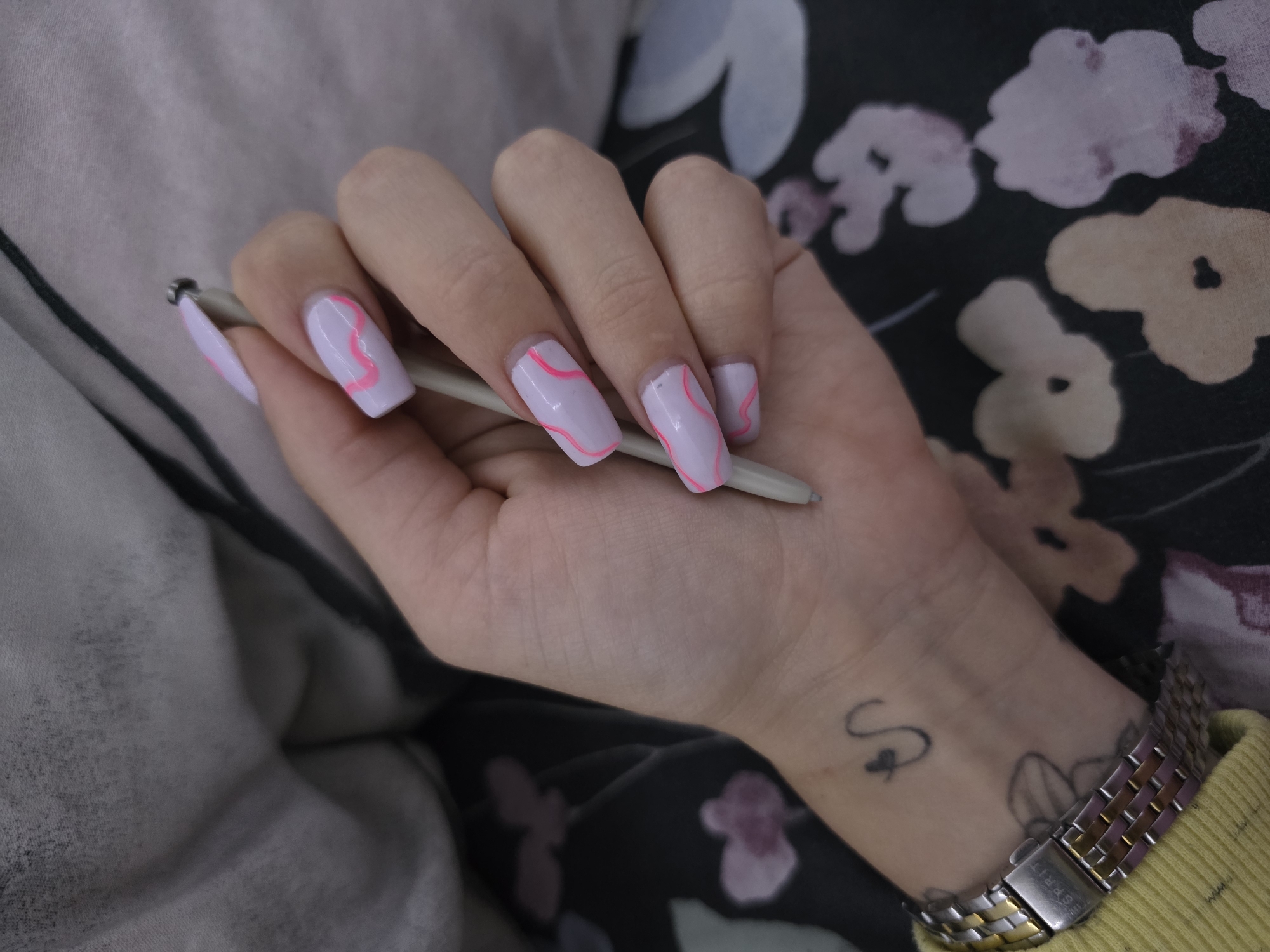 The image presents a close-up view of a person's hand holding a stylus or pen, set indoors. The hand, likely female, is positioned in the center, with fingers gently gripping a slender, light gray stylus. The nails are long and squared, painted with a light lavender or pale purple polish, and each visible nail features a distinct bright pink wavy line pattern.
On the wrist, a metallic wristwatch with a two-tone link band (silver and a rose gold or gold hue) is visible. The brand name "ESPRIT" is clearly etched on the clasp. Above the watch, on the back of the hand, there is a small black tattoo depicting a stylized cursive "S" with a small heart or loop. Part of another, larger tattoo with an abstract or floral design is partially visible beneath the watch. A yellow ribbed fabric, likely a sweater cuff or sleeve, is seen near the watch.
The background comprises soft fabrics. To the upper left, a light gray or off-white textured fabric is present. To the right and below the hand, a darker fabric, possibly black or dark green, displays an abstract floral pattern in shades of light pink, beige, and white. The setting appears casual and indoor. The provided location context for this media file is Babovo, Bulgaria.