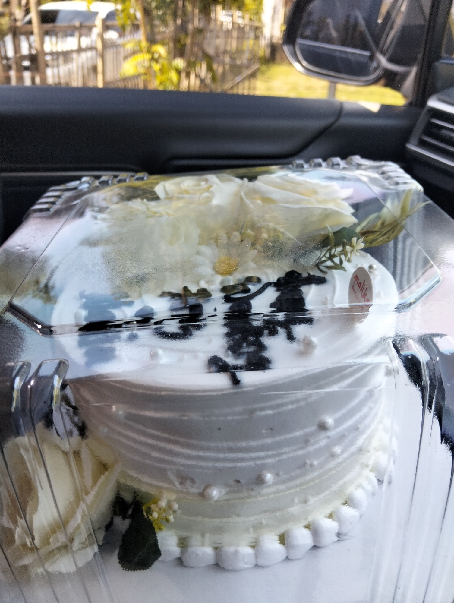 An overhead, slightly angled, close-up shot shows a white cake inside a clear plastic container. The cake is decorated with white frosting and appears to be adorned with white roses, white daisies, and greenery. There are also small pearl-like decorations on the side of the cake. On the top of the cake, there are black, abstract icing decorations. A small, circular sticker with red text and a red outline, possibly a brand logo, is visible on the right side of the cake. The cake is situated inside a car, with the dark interior of the vehicle visible in the foreground and background. The outdoor environment, including trees and a fence, can be seen through the car window. There are no people visible in the image. The image depicts a cake that is likely being transported. The location is identified as Cottrueng, Indonesia.