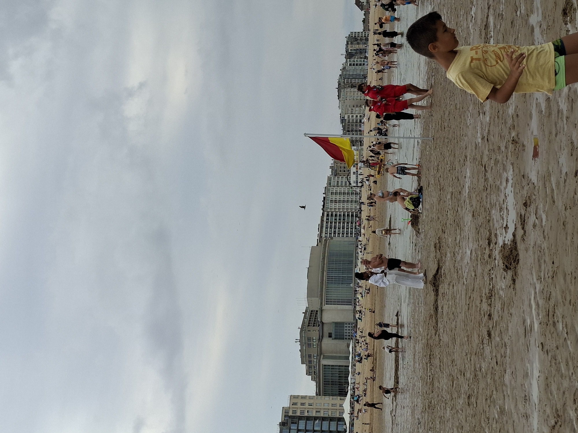 The image depicts a busy beach in Ostend, Belgium, under an overcast sky. Numerous people are scattered across the sandy shore, engaging in various activities such as wading in the shallow water, playing, and conversing. In the foreground, a young boy in a yellow t-shirt and green shorts walks towards the right side of the frame, looking down. A lifeguard flag, red and yellow, is prominently displayed on a pole near the center of the image. Behind the beachgoers, a long row of multi-story buildings lines the waterfront, characteristic of a coastal city. The sand appears wet and reflective in places, indicating recent tides or shallow water. The overall atmosphere suggests a moderately active beach day, despite the cloudy weather.