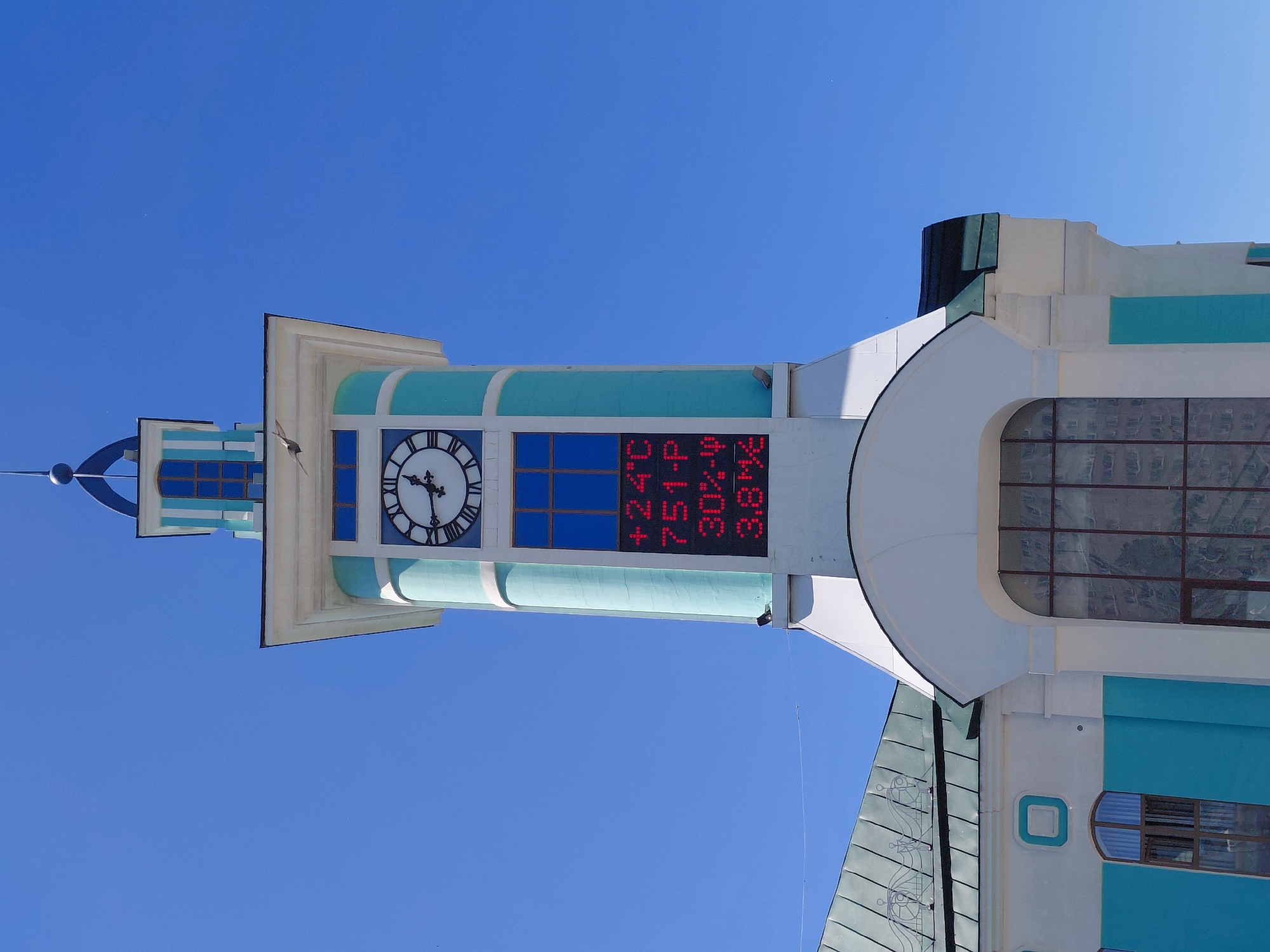 An eye-level, full shot captures a clock tower against a clear, bright blue sky. The tower, predominantly white and turquoise, features a circular clock with Roman numerals and a digital display below that shows "+24C 751-P 30% 3.8M". A bird is in mid-flight near the top of the tower. The tower is attached to a building with a green, corrugated roof. The lower portion of the building is painted white and turquoise, with arched windows. The scene appears to be in an urban environment, though no specific landmarks are identifiable.