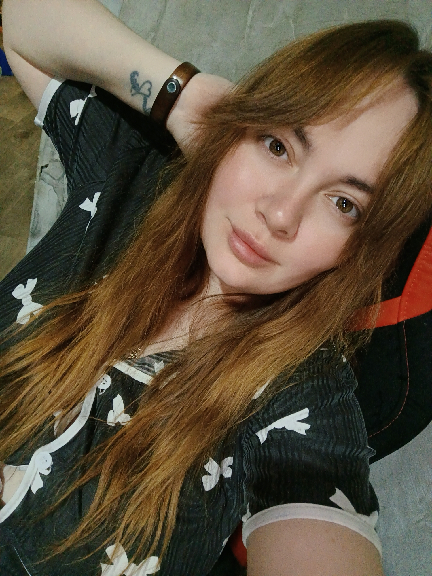 A young woman with long, reddish-brown hair and brown eyes is featured in a close-up, upward-angled photo, likely a selfie. She has a subtle, calm smile and looks directly into the camera. Her hair is parted, with bangs framing her face.
She is wearing a dark-colored top, possibly black or dark grey, adorned with a white, abstract pattern resembling stylized anchors or bows. The top has a V-neckline with white buttons and appears to be made of a textured fabric. A delicate chain necklace with a small pendant is visible around her neck. Her right arm is raised and rests behind her head, revealing a faded black tattoo on her inner forearm, which appears to be a heart or stylized bow design. A brown leather or wooden bracelet with a metallic stud is worn on her right wrist.
The setting is indoors. A textured grey surface, possibly a wall or floor, is visible in the background to the left. To her right, part of a black and red gaming or office chair can be seen. The lighting is soft and even. There is no discernible text visible in the image. The location context provided for this image is Chaikovsky, Russia.