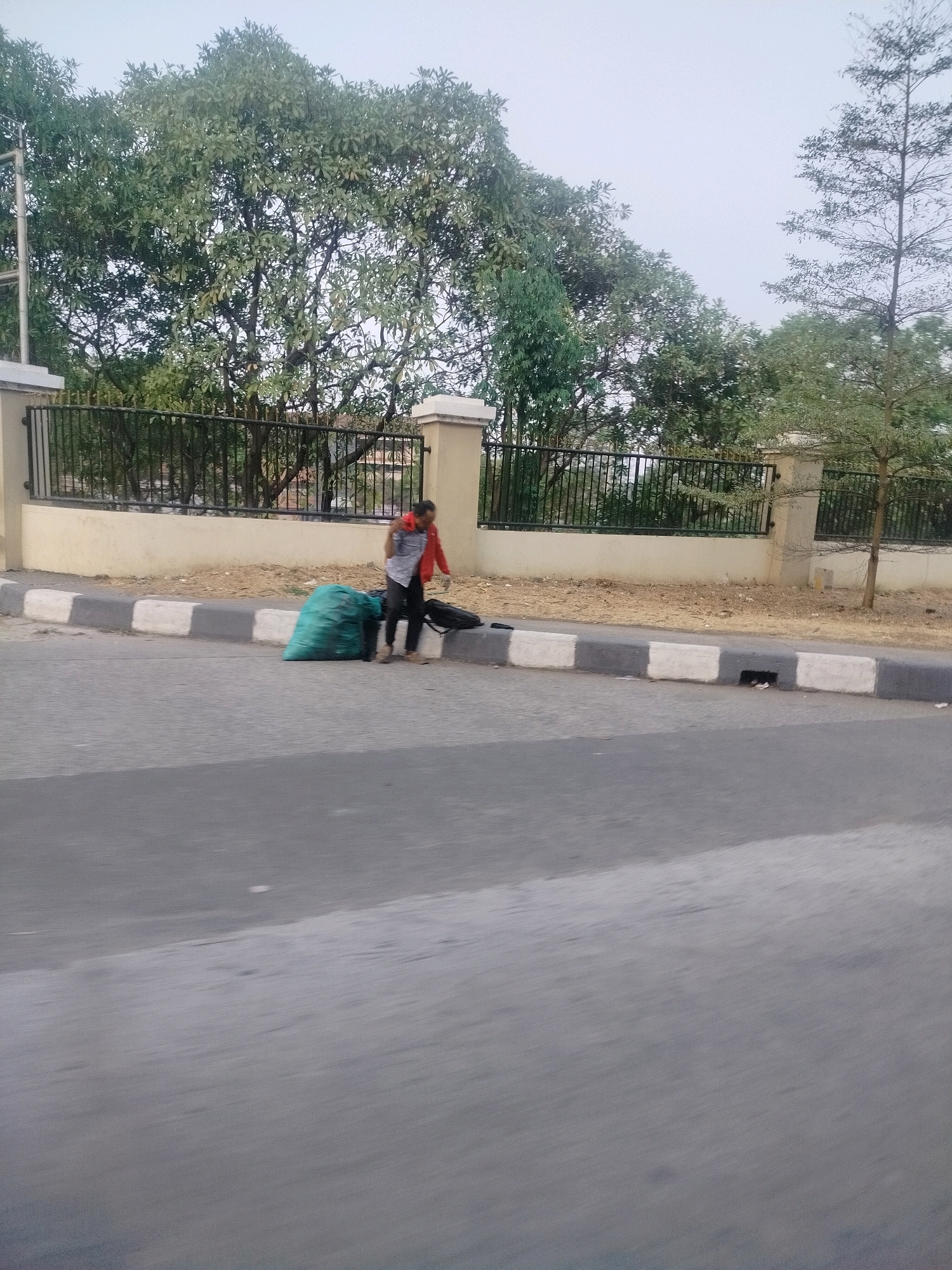 A person wearing a red jacket and black pants is standing on a sidewalk next to a large green trash bag and a black bag. The person appears to be adjusting the red jacket. The sidewalk has a pattern of white and gray blocks. Behind the sidewalk is a beige wall with a black metal fence. Trees are visible behind the fence. The image is taken from a moving vehicle, indicated by the motion blur in the foreground of the asphalt road. The location is the city of East Jakarta, Indonesia.