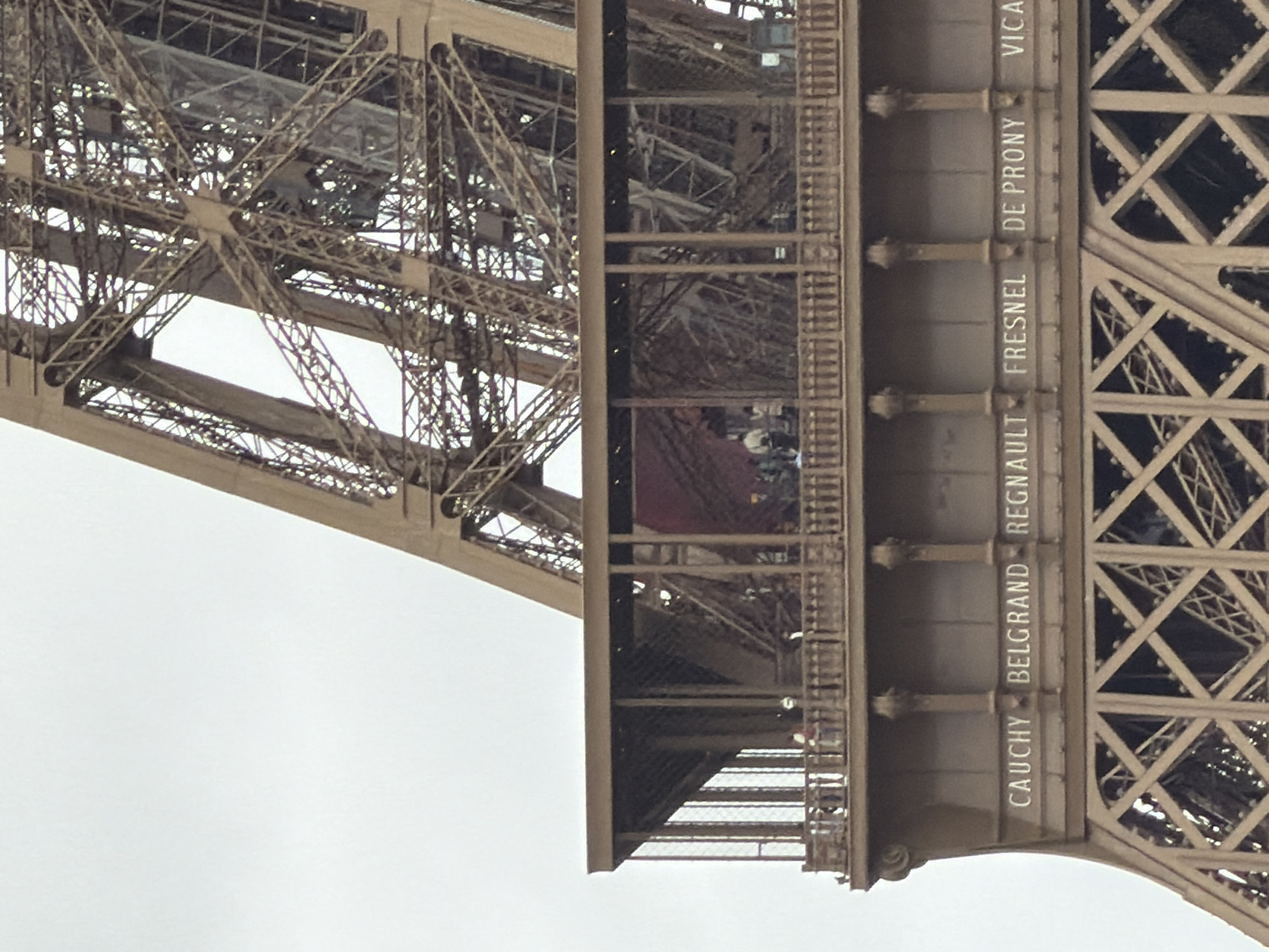 This image provides an upward close-up view of a section of the Eiffel Tower in Paris, France, against a bright, likely overcast sky during the daytime. The perspective emphasizes the intricate wrought-iron latticework and massive structural beams that form the iconic monument.
On the right side of the frame, a vertical support beam of the tower is prominently displayed. Engraved into its surface are several names of notable French scientists, engineers, and mathematicians, a tribute to their contributions. Visible names, from bottom to top, include "CAUCHY", "BELGRAND", "REGNAULT", "FRESNEL", "DE PRONY", and partially visible "VICA" (likely VICAT). Each name is accompanied by decorative architectural elements below it.
Within the complex metallic framework, a platform or intermediate level is discernible. Small figures, likely tourists or visitors, are visible on this platform, suggesting the typical activity of sightseeing and movement between the tower's different tiers. Railings are also visible along the edges of the platform. The image captures both the monumental scale and the fine details of the Eiffel Tower's engineering and historical commemoration.