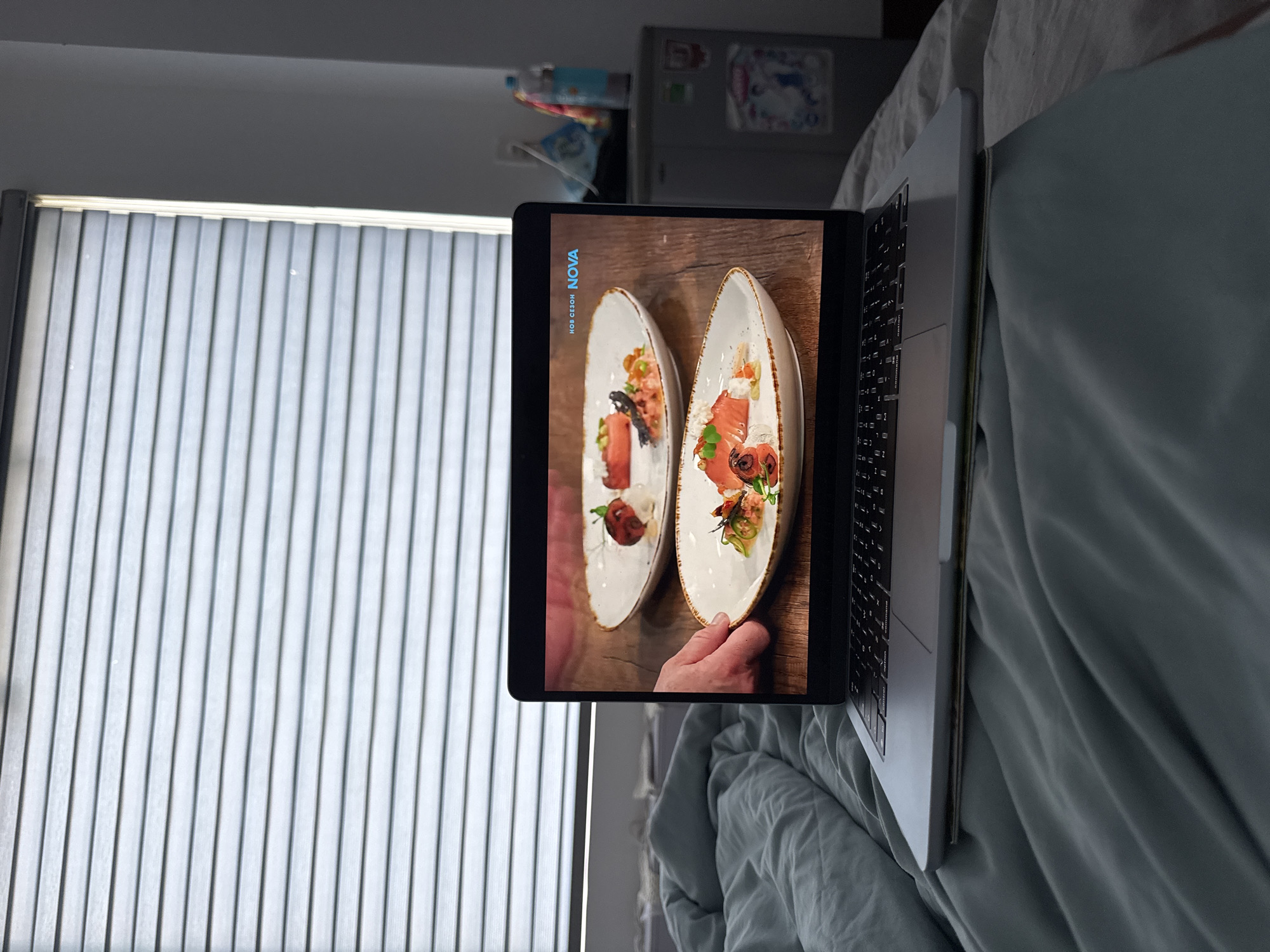 The image shows a laptop resting on a bed with light blue bedding. The screen displays a video of food presentation, specifically two plates with salmon-based dishes. The background includes a window with closed blinds and a refrigerator unit. The location is implied to be a residential setting, potentially in the city of Đà Nẵng, Vietnam based on the video shown in the laptop. A hand can be seen holding the plates.