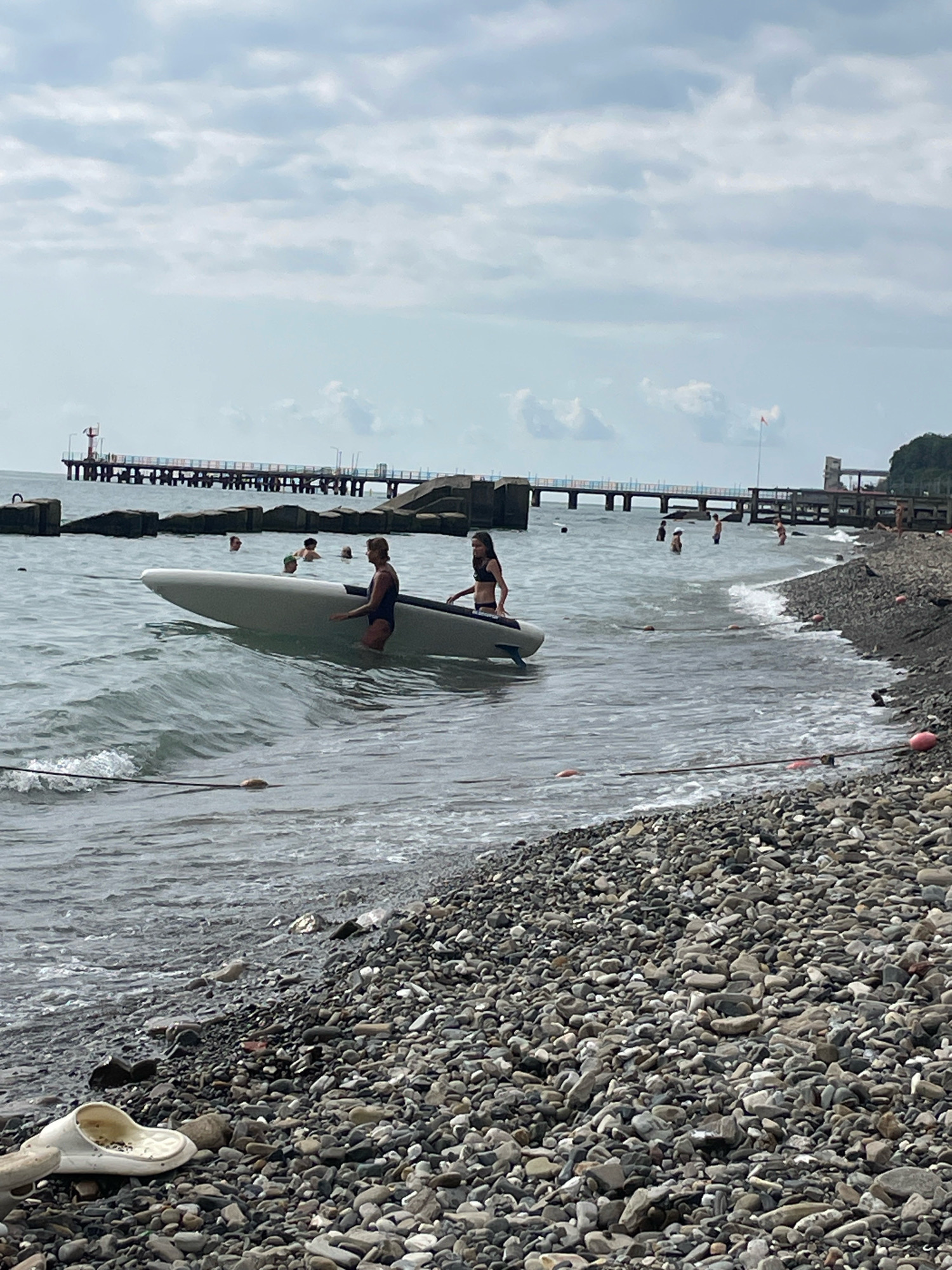 An overcast day at a pebble beach in Sochi, Russia, where two individuals are partially submerged in the sea, pushing a white surfboard towards the shore. The water laps gently against the beach, and in the background, a long, industrial pier stretches out into the sea, with several people visible on it. The sky is filled with a layer of grey clouds.