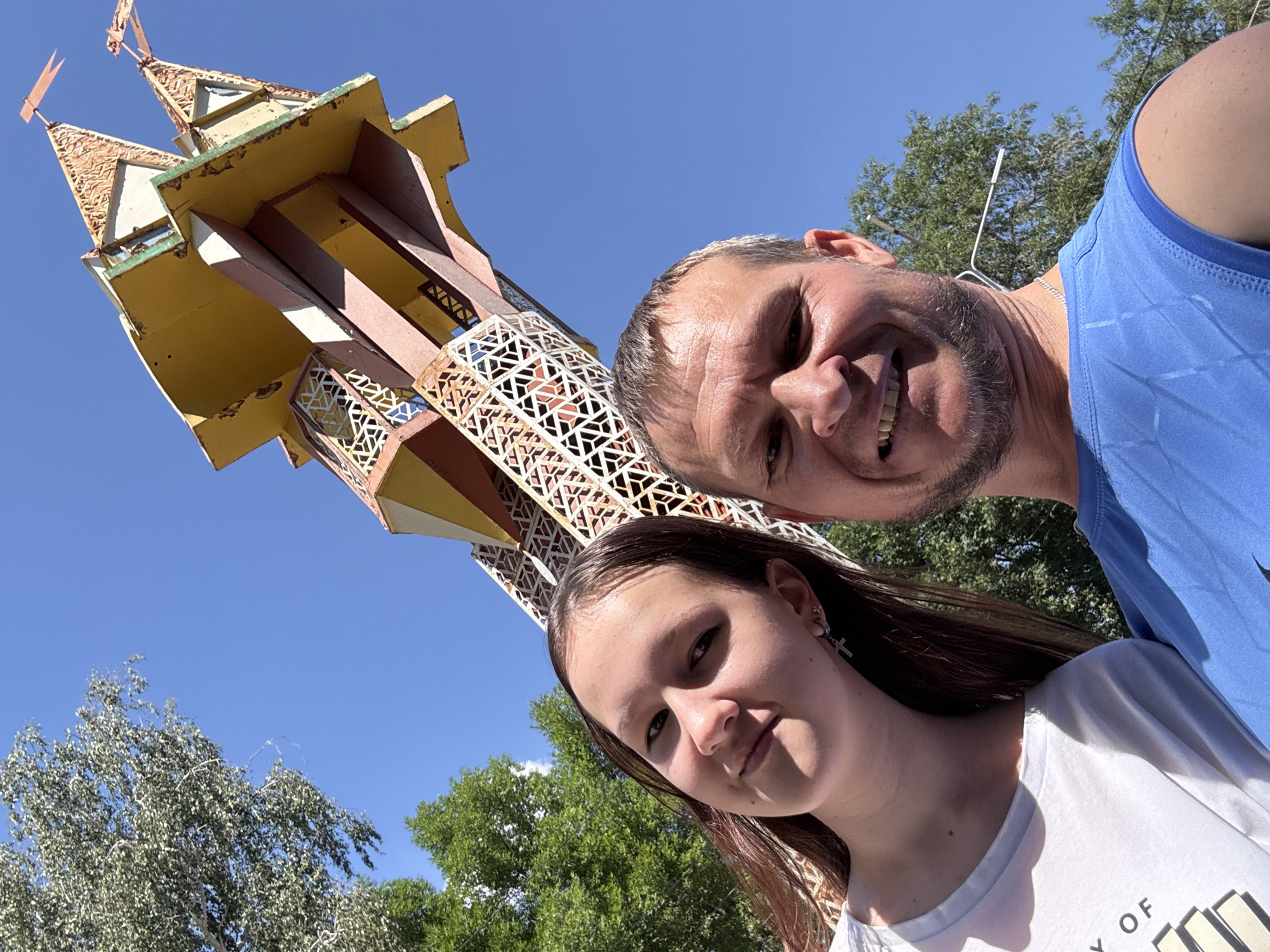 In this selfie, a man and a young woman are positioned in the foreground, looking towards the camera. They are outdoors, under a clear blue sky. Behind them, a distinctive, colorful structure resembling a stylized castle or tower stands tall. The structure is made of various materials and features intricate latticework and pointed rooftops with small flags. Green foliage from trees is visible on the left and right sides of the image, suggesting a park or outdoor recreational area. The lighting indicates it is daytime. The image appears to have been taken in Oskemen, Kazakhstan, possibly near a playground or amusement area, given the nature of the structure in the background.