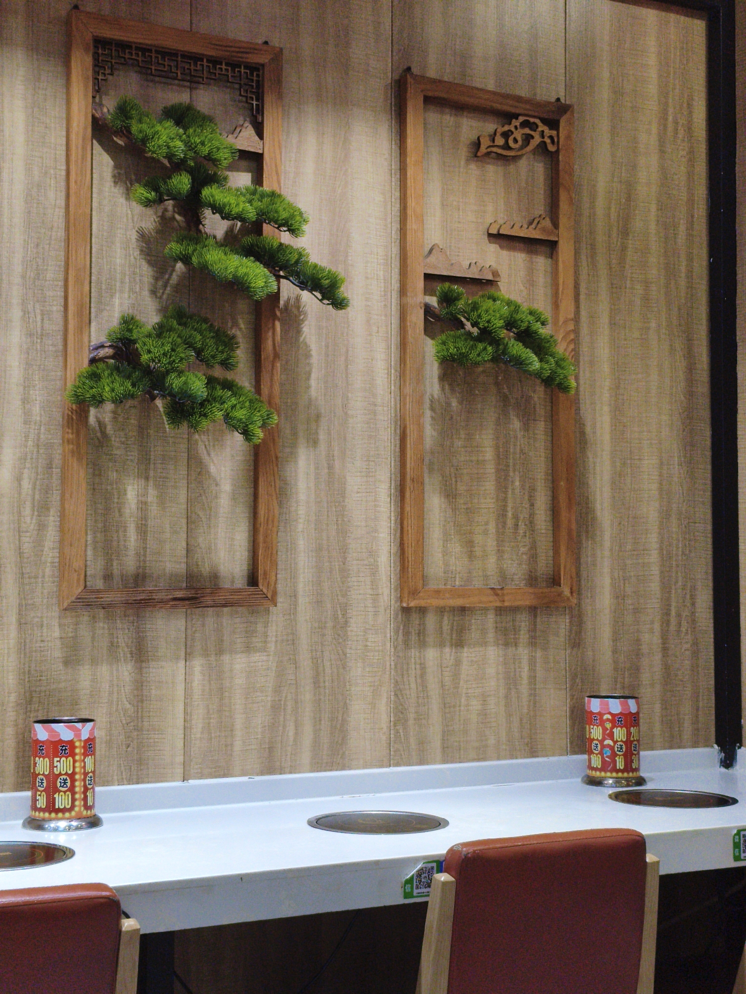 The image depicts a dimly lit interior space, likely a restaurant, with a wood-paneled wall as the backdrop. Two framed decorative elements, resembling traditional Chinese window grilles with faux pine branches, are mounted on the wall. In the foreground, a white countertop is visible, featuring two circular induction cooktops. On the left side of the countertop sits a cylindrical red and white container with Chinese characters and numbers, possibly indicating a promotion or pricing. To the right, another similar container is present. Two red chairs with wooden frames are partially visible in the foreground. The overall aesthetic suggests a contemporary Asian dining establishment. Without additional information, it is impossible to determine the specific location in Mudanjiang, China.
