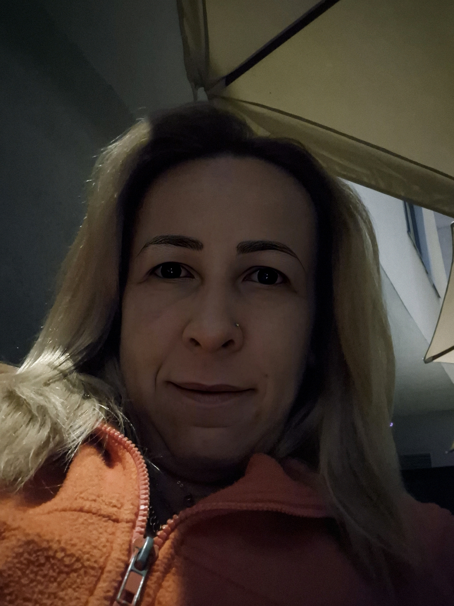 An indoor selfie shows a woman from the chest up, wearing an orange hooded jacket. She has blonde hair and a nose ring, and is looking at the camera with a slight smile. The background is dimly lit, with hints of furniture and a ceiling fan visible. The lighting suggests it is nighttime or a poorly lit indoor space.
