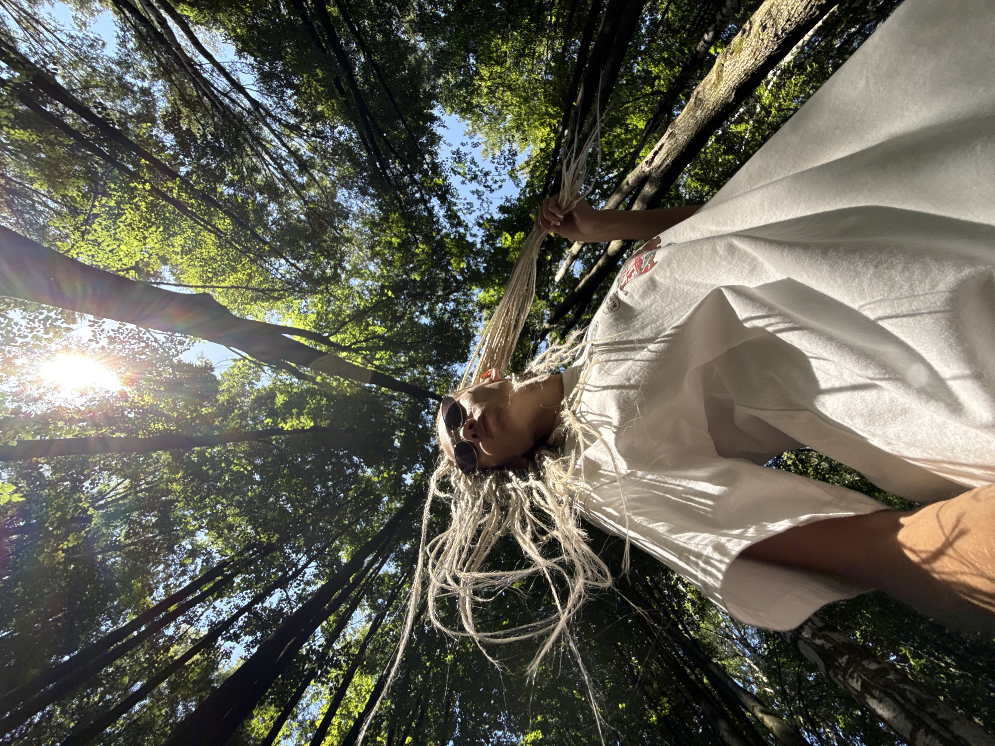 In the city of Rusanovo, Russia, a low-angle shot captures a person from below, looking up into a canopy of trees. The sun shines brightly through the leaves, casting dappled light and lens flare across the scene. The person, wearing sunglasses and a white t-shirt adorned with a graphic, has long, light-colored dreadlocks that are being held and appear to be in motion. The trees are dense, with branches and foliage creating a natural ceiling overhead. The perspective emphasizes the height of the trees and the vastness of the sky peeking through.