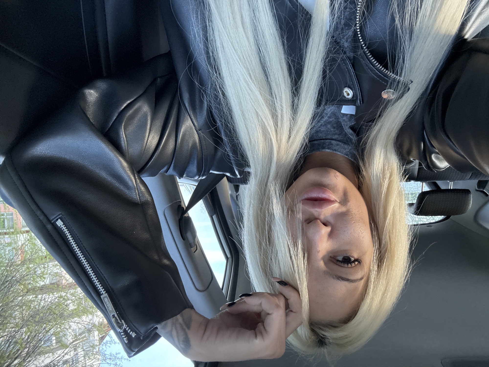 A person is in the front seat of a vehicle. The person is wearing a black leather jacket and a gray shirt. The person has long, blonde hair. The person is holding their hand up to their face. The vehicle is located in an urban environment. Trees and buildings are visible through the window. The exact location is unknown.