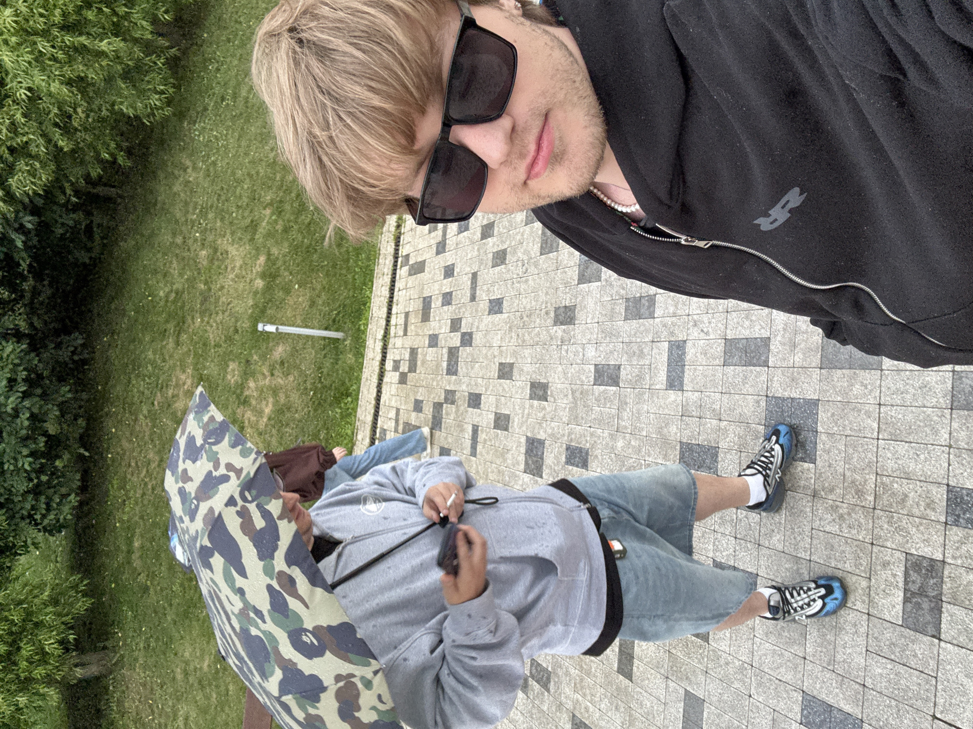 Two people are standing on a paved area next to a grassy lawn with trees. The person in the foreground is wearing sunglasses, a black zip-up hoodie with a logo, and pearl necklace. This person is holding a phone or camera and taking a selfie. Their hair is blonde and short. The second person is partially visible, holding a camouflage umbrella. They are wearing a grey hoodie and denim shorts. The ground is tiled with a pattern of grey, white, and black squares. The scene appears to be outdoors, possibly a park or a residential area, in Khimki, Russia. The presence of the umbrella might indicate it is raining or has recently rained. The lighting suggests it is daytime.