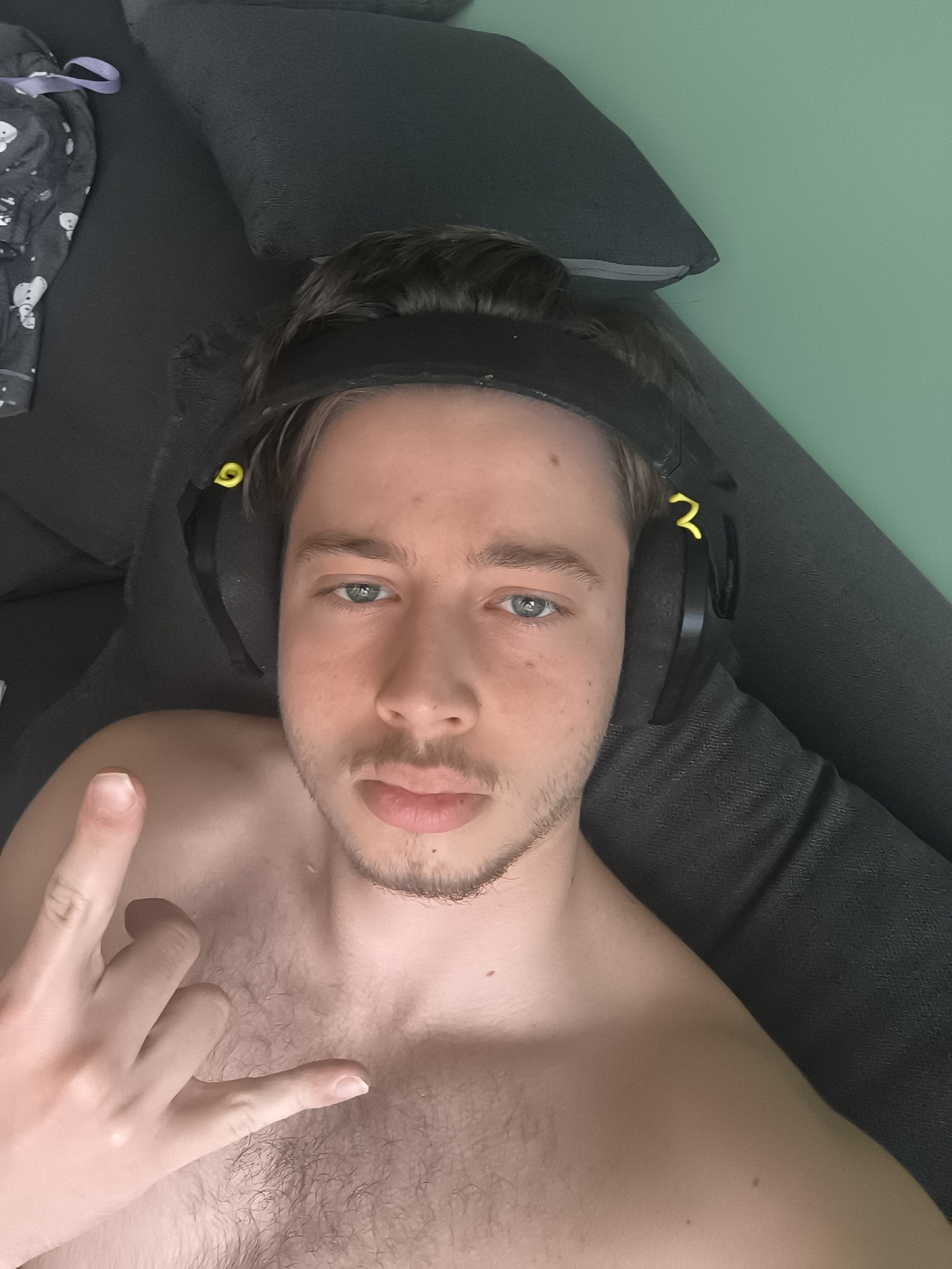 A young man takes a selfie while lounging indoors, looking directly at the camera with a neutral expression. He has fair skin, light brown hair, and blue eyes, with light stubble on his face and some chest hair. He is shirtless and wears black over-ear headphones with yellow accents, which are pushed back onto his head. His left hand is raised, performing a "rock on" hand gesture. The background features a dark gray upholstered surface, likely a sofa or bed, on which he is resting. To the upper left, a piece of patterned pajama fabric with white snowman figures and a purple drawstring is visible. A light green wall is visible in the upper right portion of the frame.