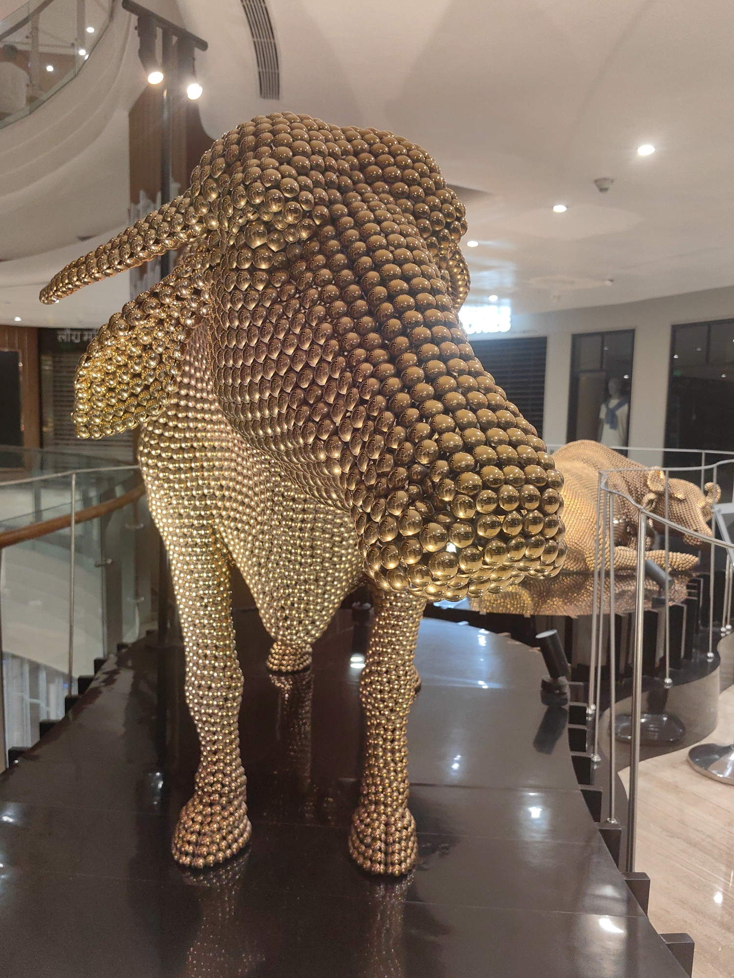A golden, metallic sculpture of a bull stands prominently in what appears to be a shopping mall, possibly in Mumbai, India. The sculpture is intricately crafted from numerous small, spherical elements, giving it a textured, scaled appearance. Its head is turned to the right, and its body is positioned to face forward. The bull is illuminated by overhead lighting, which creates reflections on its metallic surface and the dark, glossy platform it rests upon. In the background, a staircase with a glass railing is visible, along with store facades and ceiling lights, typical of a commercial indoor environment. A second, partially visible bull sculpture made of similar material is situated behind the main subject. The overall scene depicts an artistic installation within a modern retail space.