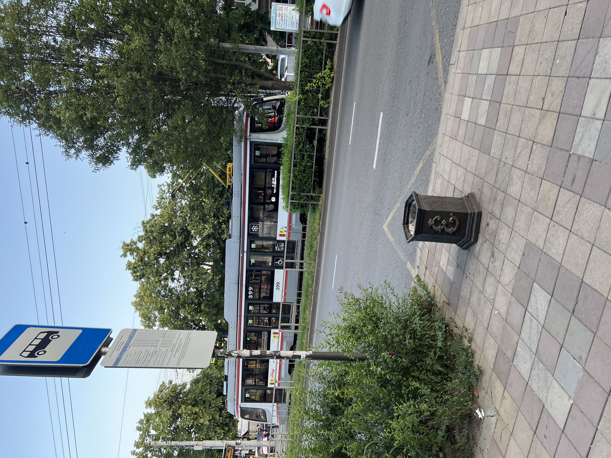 In Krasnodar, Russia, a street scene unfolds with a tram stopped at a bus stop. The tram, numbered 399, has its doors open, and passengers are visible inside. A bus stop sign is prominently displayed, indicating it's a public transportation hub. The tram is on a paved road, bordered by a sidewalk made of patterned tiles. A metal fence separates the sidewalk from the street, and behind the fence, lush green bushes and trees line the area, suggesting a well-maintained urban environment. A black trash receptacle with decorative metalwork stands on the sidewalk. The sky above is clear and blue, hinting at fair weather. The overall impression is a snapshot of daily life in a Russian city, focusing on public transit and its surroundings.