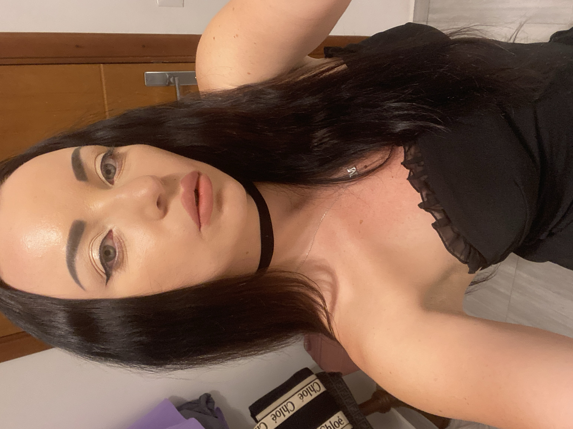 The image shows a close-up, waist-up portrait of a woman with long, dark hair and fair skin. She is wearing a black strapless top with a ruffled neckline and a black choker necklace. She has winged eyeliner and pink lipstick. A silver necklace with a letter "M" pendant is visible. She is leaning slightly to her right. In the background, to the left, is a chair with a bag on it that appears to have "Chloe" written on it. To the right, there is a wooden door with a silver handle and a white wall. The lighting suggests an indoor setting. The scene is likely in a residential area or hotel in Sharm El Sheikh, Egypt, given the woman's appearance and the warm tones of the background, common in Egyptian architecture.
