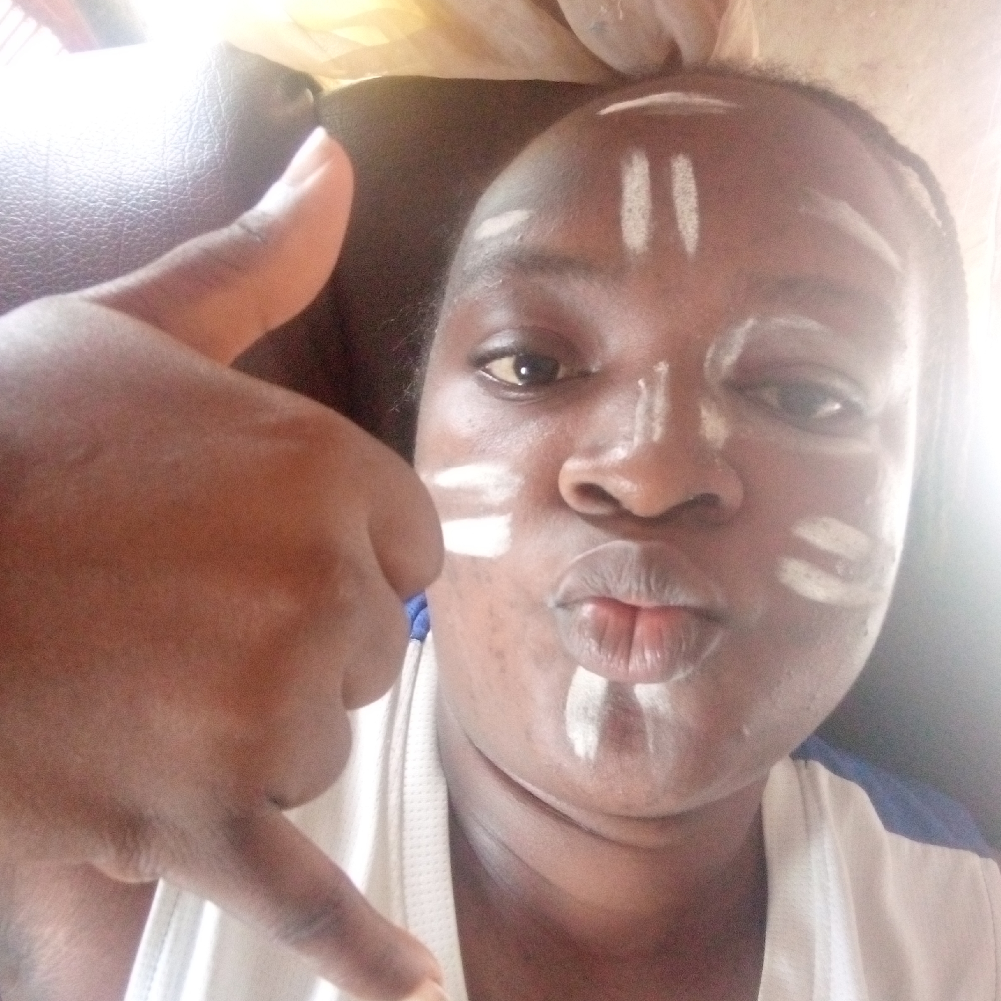 A close-up selfie depicts an individual with dark skin, adorned with white facial markings. Two prominent vertical white lines are visible on the forehead, with single horizontal lines under each eye, a vertical line on the bridge of the nose, and another on the chin. The person is looking directly at the camera with their right eye and has their lips slightly puckered. In the foreground, a left hand is raised, making a "shaka" or "hang loose" gesture (thumb and pinky extended).
The individual is wearing a white garment, possibly a t-shirt, with a hint of blue fabric visible on the upper left. The background consists of a dark, textured surface, likely furniture upholstery, brightly lit by natural light streaming from the top left, suggesting a daytime setting. The facial markings, in the context of Ogidi, Nigeria, likely indicate participation in a cultural event, ceremony, or celebration. The overall expression and hand gesture convey a playful or confident mood. No text is visible in the image.