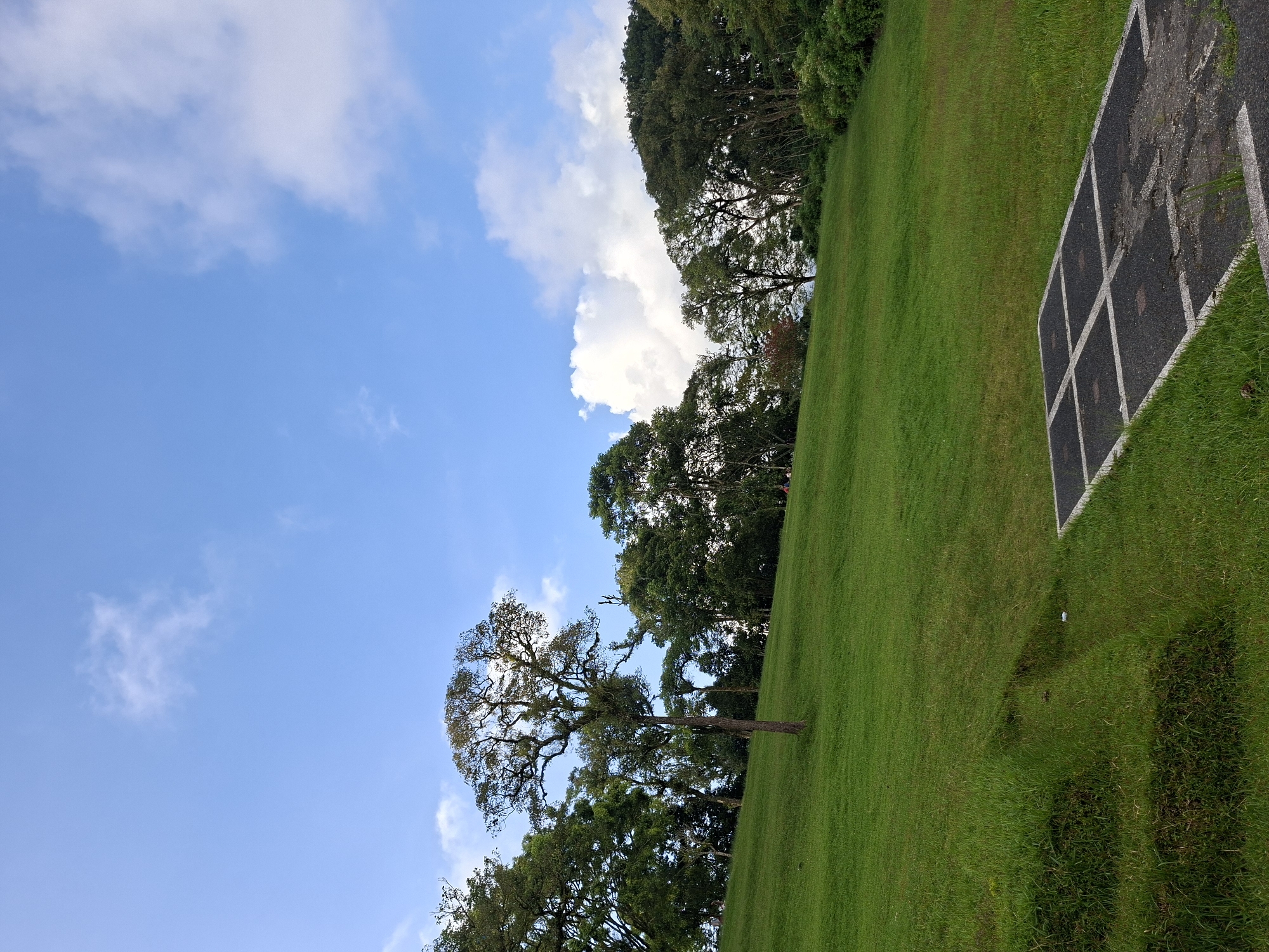 The image depicts a landscape in Baturiti, Indonesia. A predominantly green lawn slopes upwards from left to right. On the right edge is a square black asphalt pad with white painted square divisions. Trees line the horizon. Above, a blue sky with cumulus clouds is visible. No people are visible.