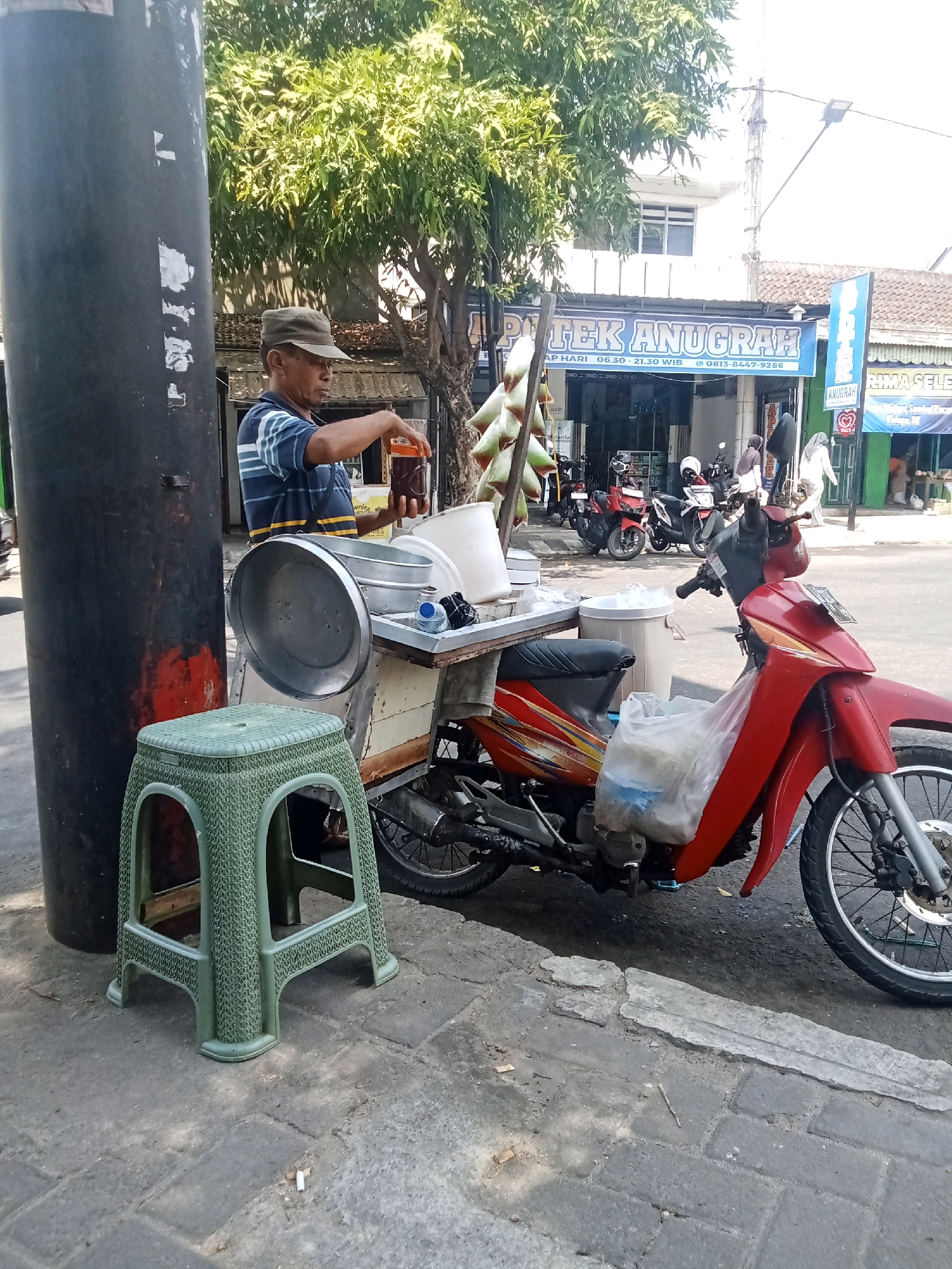 The image depicts a street scene in Sukowinangun, Indonesia, where a man is preparing a beverage. He stands next to a mobile cart attached to a red motorcycle, holding an orange-lidded glass jar filled with a dark liquid. A green plastic stool is positioned to his left. The cart itself is laden with various containers, including a large metal bowl and a white plastic tub. In the background, a pharmacy named "APOTEK ANUGRAH" is visible, along with other storefronts and parked motorcycles. The street is paved with interlocking bricks, and a large dark pole stands on the left side of the frame. Sunlight casts distinct shadows, indicating it is daytime.