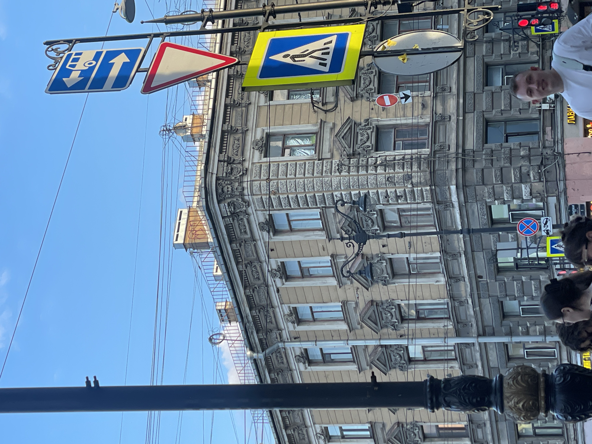 This image captures a vibrant street scene in Saint Petersburg, Russia, under a clear blue sky on a sunny day. The perspective is tilted, showcasing ornate historical buildings that line the street. The building on the left features a lighter, smoother facade, while the one on the right displays a more textured, block-like exterior, both adorned with intricate architectural details around windows and cornices.
Numerous overhead wires crisscross the sky, indicative of active urban infrastructure. Several street poles are prominent, bearing various traffic signs. These include a blue rectangular sign with a white upward arrow and a downward tram or bus symbol, a red-bordered triangular sign, and a distinctive square yellow sign with a blue triangle containing a white pedestrian crossing symbol. Other signs visible are a red circular 'No entry' sign with a white horizontal bar and a right arrow, and a blue circular 'No stopping' sign with a red 'X'. A traffic light showing red is also present.
In the foreground, a dark, decorative street pole stands. Further down the street, a person in a white shirt is partially visible, looking towards the camera with a neutral to slightly positive expression. A group of other people can be seen further away, suggesting general pedestrian activity. A convex mirror is attached to one of the street poles. On a building in the distance, an external fire escape or access ladder is visible. Partial Russian text, "ЛАВК...", likely indicating "shop," is visible on a sign on one of the buildings. The scene depicts a typical busy yet charming urban environment.