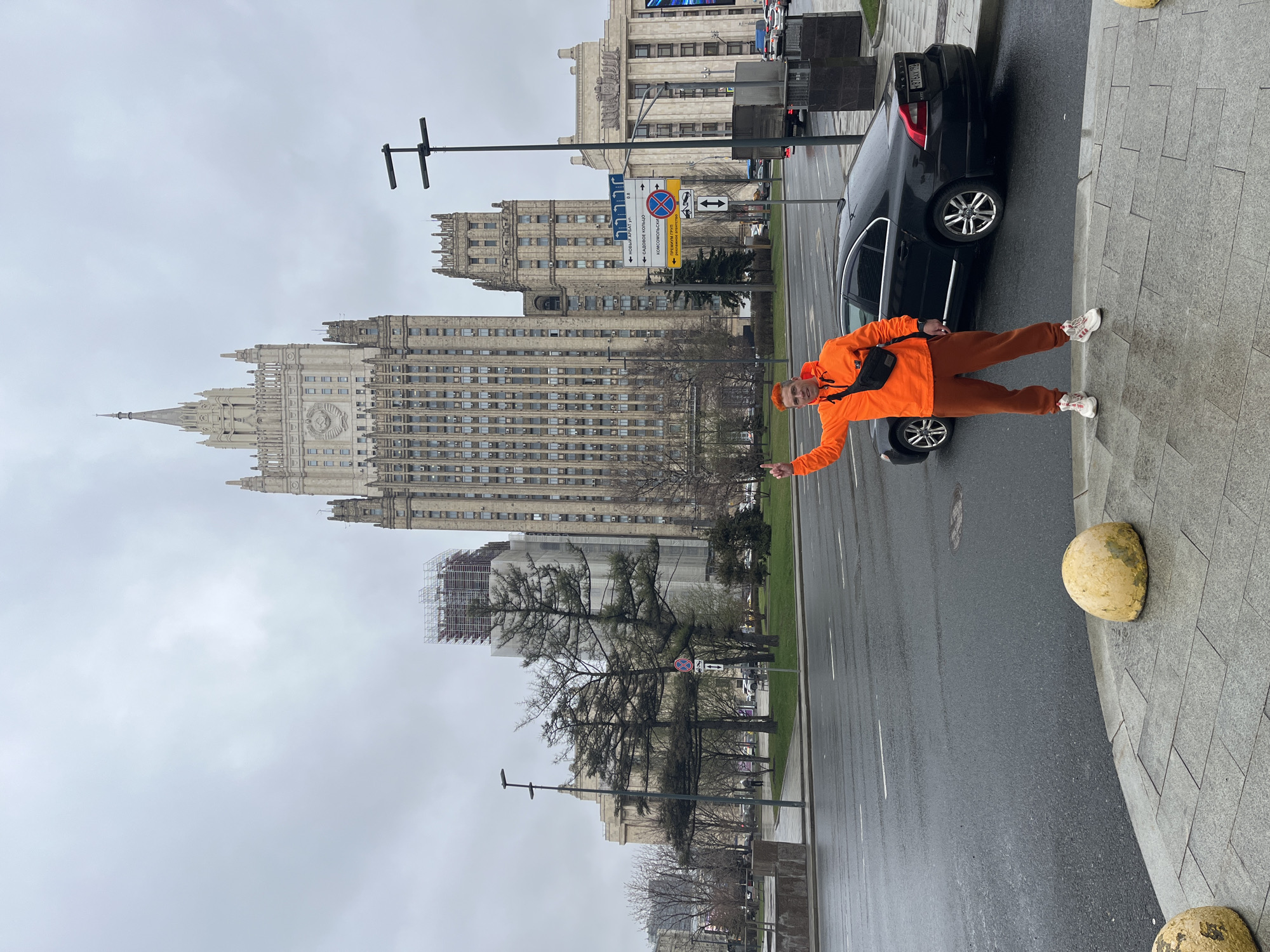 The image shows a man in an orange jacket and pants standing on a sidewalk in Moscow, Russia. He is pointing towards the Ministry of Foreign Affairs building, a Stalinist skyscraper. A black car is parked beside him on the street, which appears wet. The sky is overcast. In the background are other buildings and street signs.