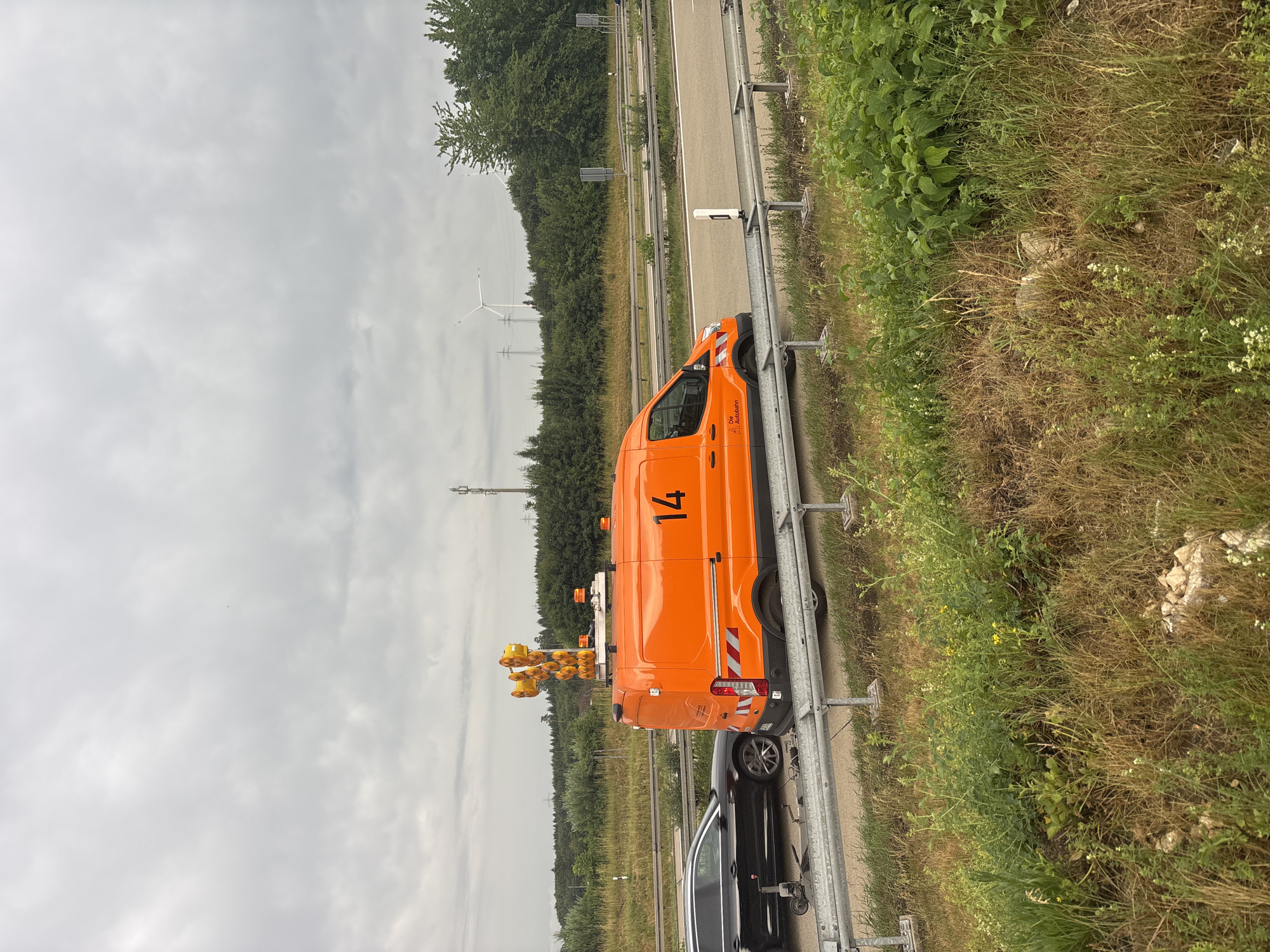 The image depicts a traffic scene on a cloudy day, likely on an Autobahn near Heidenheim an der Brenz, Germany. A bright orange maintenance van, identified by the number "14" and a logo that includes "Die Autobahn," is parked on the side of the road. The van is equipped with a tall array of warning lights and what appears to be a camera or sensor unit mounted on top. Adjacent to the van is a black car, possibly disabled, with a trailer hitch. Both vehicles are positioned behind a metal guardrail. The surrounding environment consists of a highway, grassy verges, trees, and in the distance, wind turbines and power lines are visible, indicative of an industrial or semi-rural area. The sky is overcast, suggesting a possibly damp or cool day. There are no visible people in the scene.