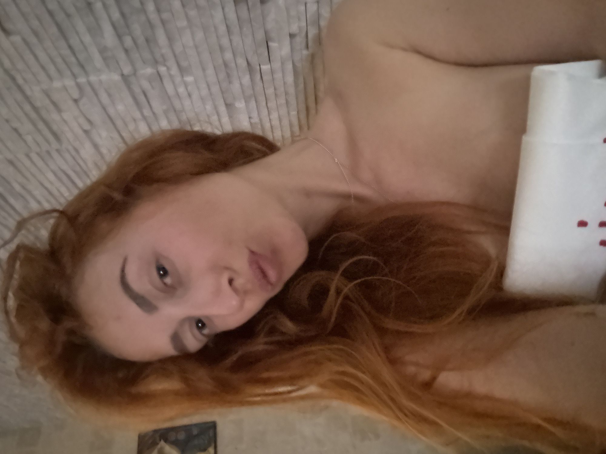 A woman with auburn hair is lying down. She is facing the camera. Her hair is spread around her head. She is wearing a necklace. A white object with red spots is visible to the right. She is lying on what appears to be a white textured surface. A portion of a decorated tile is visible in the bottom left corner. Location details are not available.