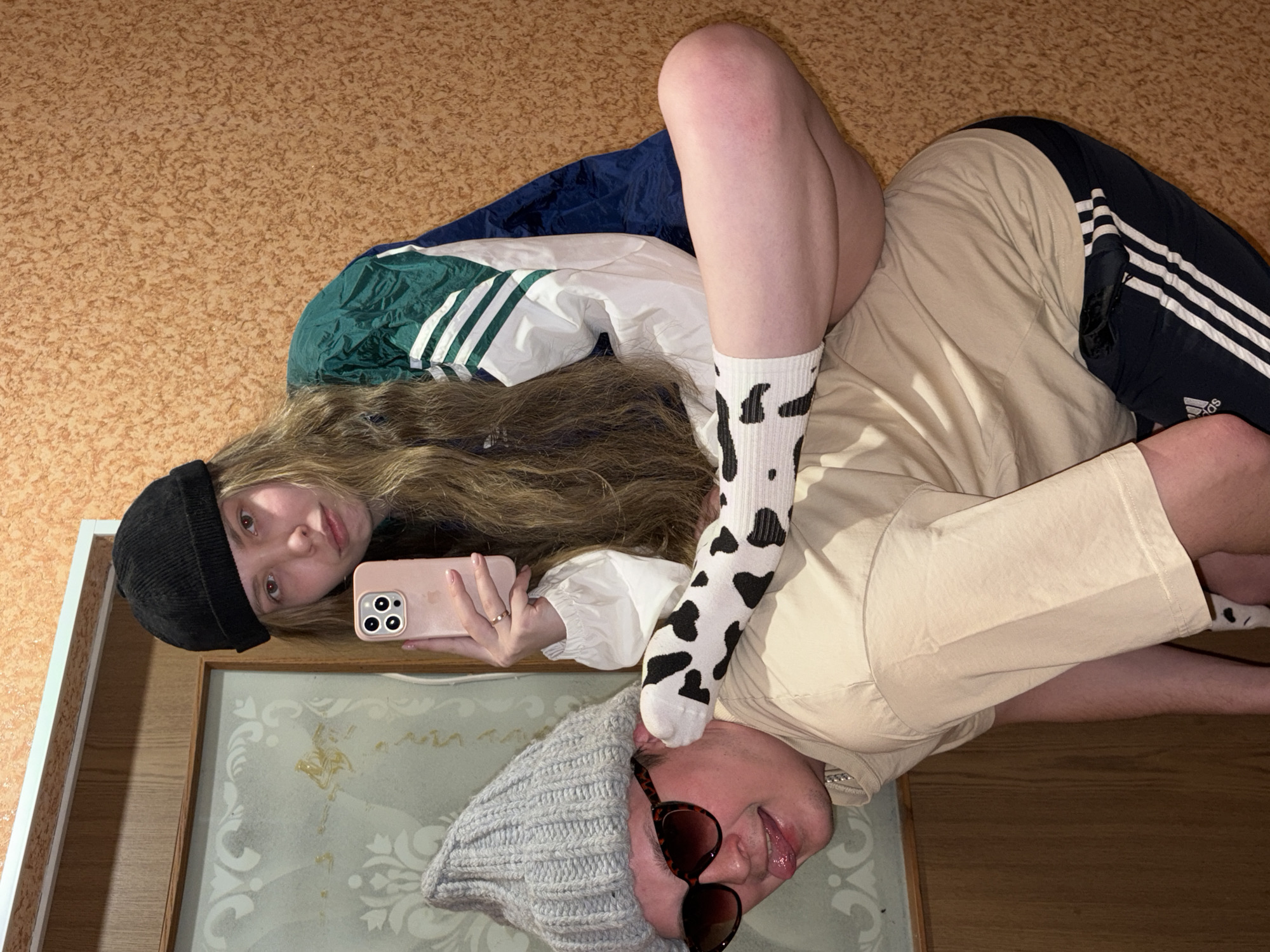 The image captures two young adults indoors, likely within a residence in the city of Chaikovsky, Russia. A female, with long wavy hair, wearing a black beanie and a green, white, and dark blue Adidas track jacket over a white long-sleeved shirt, is positioned on the back of a male. Her left leg, adorned with a white sock featuring black cow-print spots, is bent with her foot resting on his back. She holds a light pink smartphone with a triple camera array in her right hand, seemingly taking a selfie, and a ring is visible on her left ring finger. The male, bent forward in a supportive posture, wears a grey knitted beanie, dark sunglasses, a light beige t-shirt, and dark blue Adidas shorts with white stripes. He displays a playful expression with his tongue extended. A silver chain is visible around his neck. The background consists of a light-colored wall with a subtle decorative pattern to the left, adjacent to a wooden frame, and an orange-brown textured wall to the right. A white frame is visible at the top left of the scene.