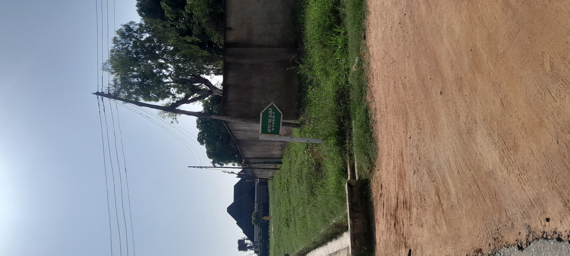 A street sign reading "JUSTICE PAUL GALUNJE STREET" is prominently displayed on a white post, pointing to the right. The sign is green with white lettering. The scene appears to be in Jalingo, Nigeria, as indicated by the presence of local architecture and vegetation. A concrete wall runs horizontally across the middle ground, separating a grassy area from the street. In the background, a large, dark-roofed building with a distinctive pyramid shape is visible, possibly a house or institutional building. Power lines are strung across the bright, clear sky, supported by utility poles. The foreground consists of a dirt road. The overall environment suggests a residential or semi-urban area. No people are clearly visible in the immediate vicinity of the sign.