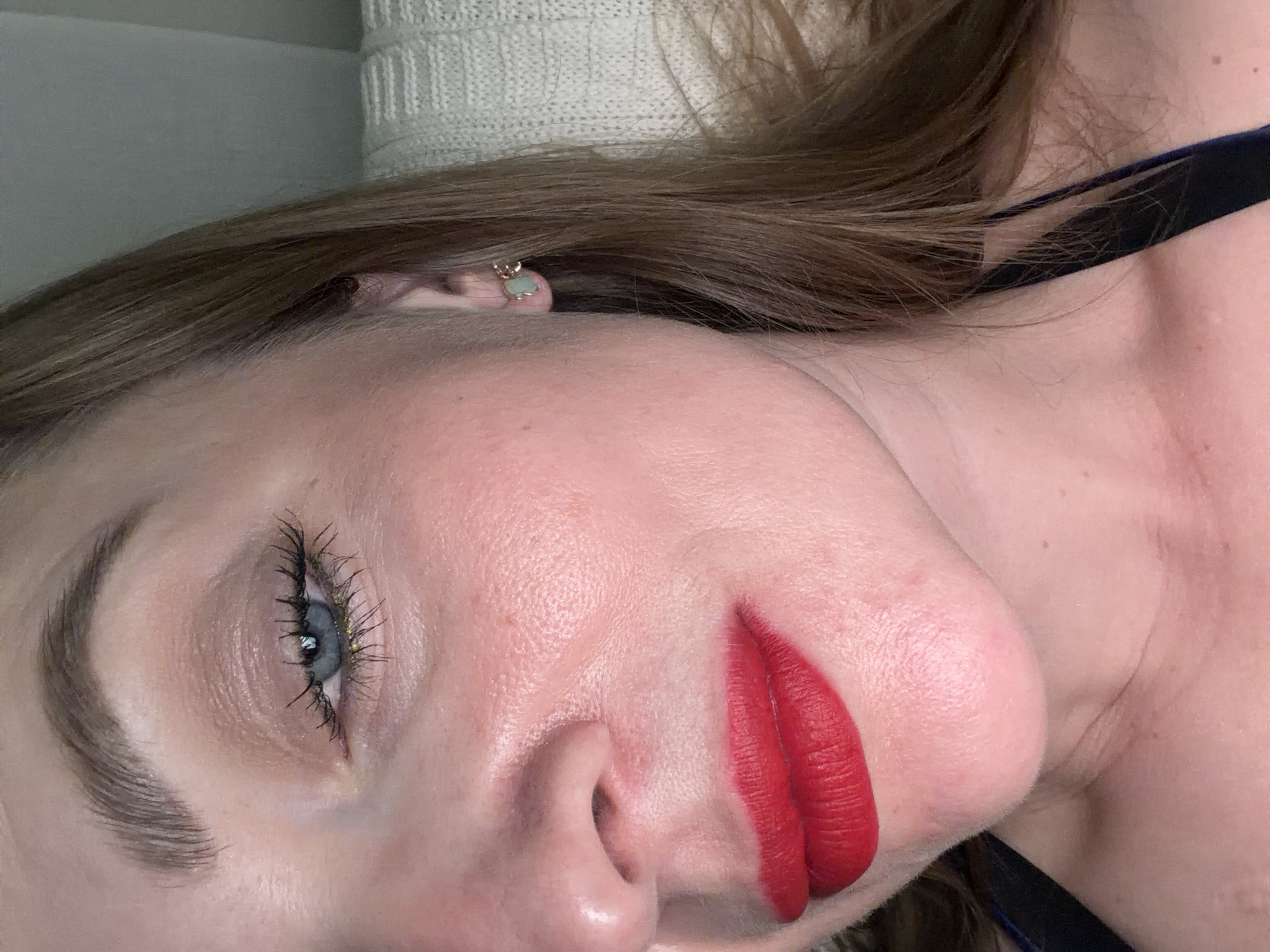 A close-up, tilted view of a person's face reveals distinct features and details. The subject's face, angled slightly, is marked by a fair complexion, with a prominent brow, long eyelashes, and a blue eye that is partially visible. The eye appears to have gold-colored eyeliner near the bottom lash line. The nose is centrally located, and bright red lipstick highlights the subject's lips. A small, light green earring is visible in the ear. The hair is light brown and partially obscures the background. The background itself consists of a creamy white knitted material and hints of a dark blue strap near the shoulder, suggesting the subject is wearing clothing. No identifiable landmarks or details indicate a specific location like Moscow, Russia. The overall impression is of a personal portrait.