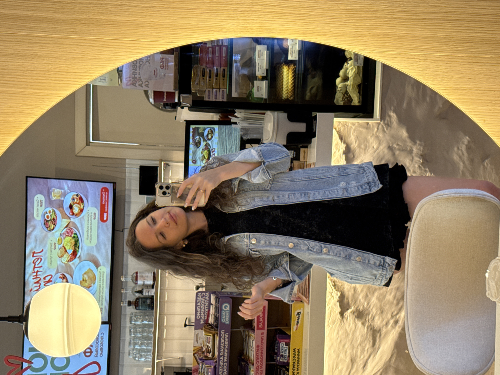 A woman is taking a selfie in a cafe. She is wearing a denim jacket over a black dress. She has long, curly brown hair and is wearing earrings and a ring. In the background, there is a digital menu displaying food items, shelves with drinks and snacks, and a display case with food items. The cafe appears to have a modern interior with a large, oval wooden mirror. The image was taken in Dolgoprudny, Russia.