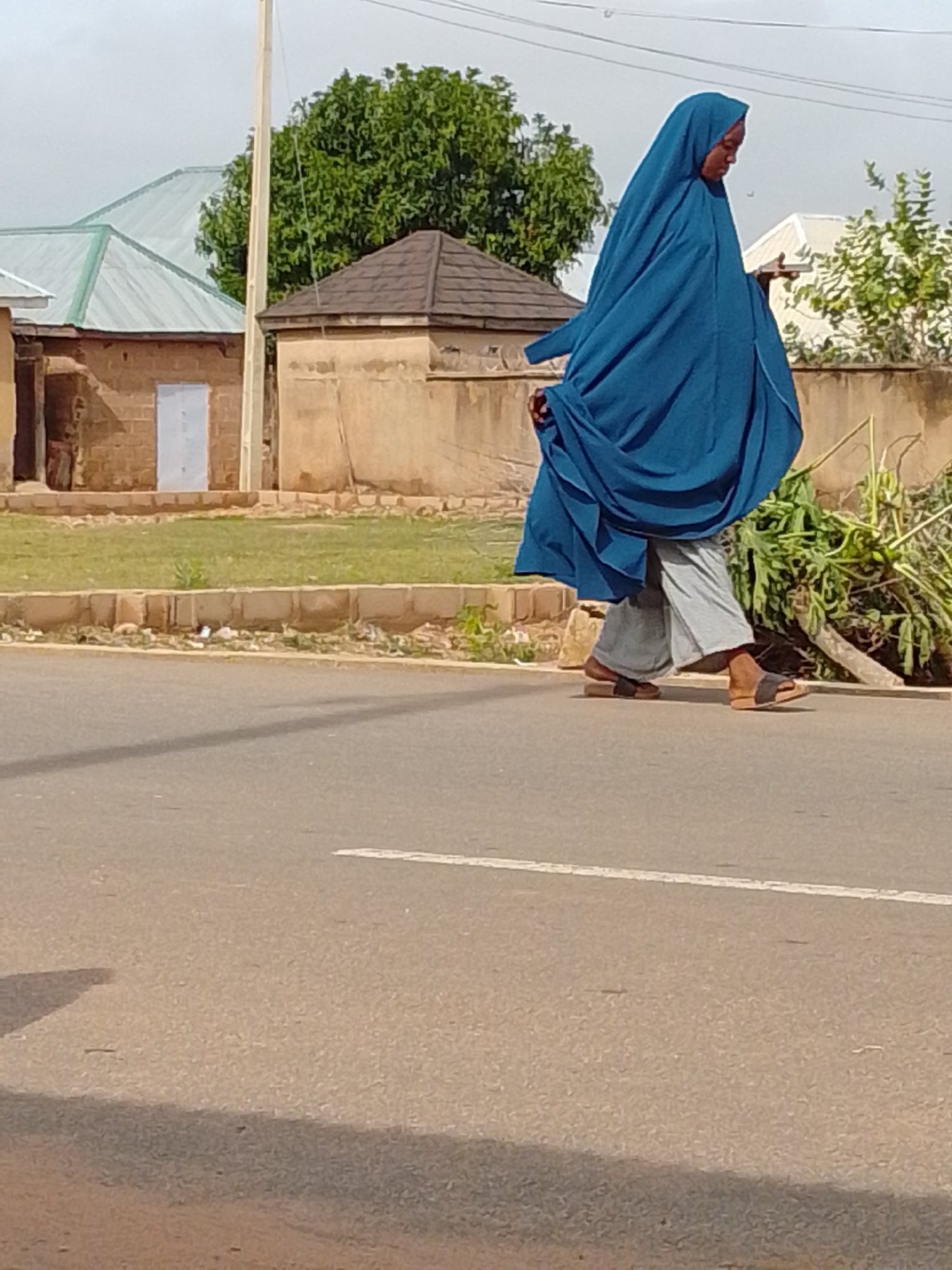 A woman wearing a blue hijab and grey pants walks on a paved road in Gombe, Nigeria. The road has a white line painted on it. To the left of the road are a grassy area and a low wall. Behind the grassy area are several buildings with brick walls and corrugated metal roofs. To the right of the woman, there is a pile of green vegetation. The sky is overcast.