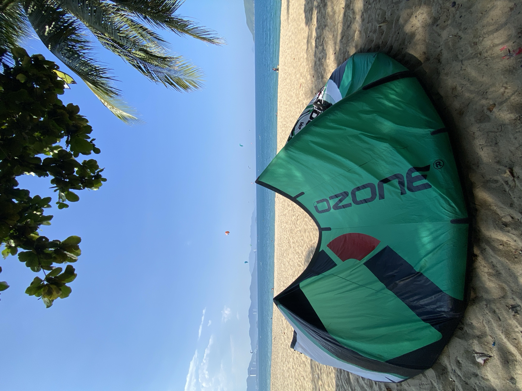 A kiteboarding kite lies folded on a sandy beach in Nha Trang, Vietnam, under a clear blue sky. The kite is predominantly green with black and white accents, featuring the "OZONE" logo in large black lettering. In the background, the calm blue ocean stretches towards a distant coastline with mountains and city buildings. Several kiteboarders are visible on the water, with some kites in the air, indicating active kiteboarding conditions. Palm fronds hang from the top right of the frame, casting shadows on the scene. The sand is golden and slightly textured with some small debris visible. The overall scene depicts a pleasant day at the beach with recreational activities.
