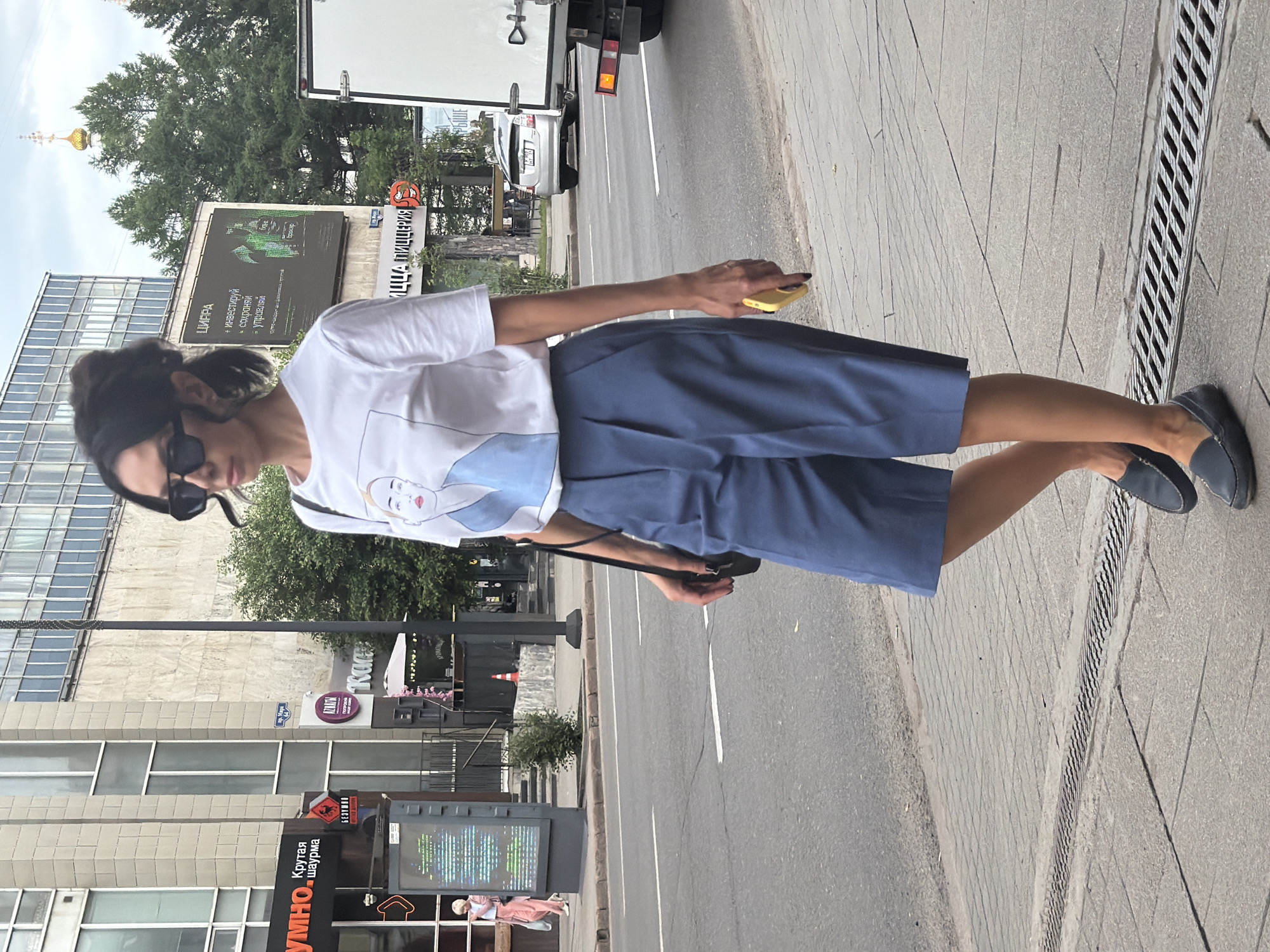 A woman walks along a sidewalk in Krasnoyarsk, Russia, on a cloudy day. She is wearing a white t-shirt with a graphic print of a woman's face and blue culottes. She has dark hair pulled back in a bun, sunglasses, and is carrying a yellow smartphone in her right hand. A black crossbody bag is slung over her left shoulder. In the background, buildings and trees line the street, with a truck visible in the middle ground. The sidewalk has a metal grate running along the edge. The scene is likely captured in the vicinity of Mira Street.