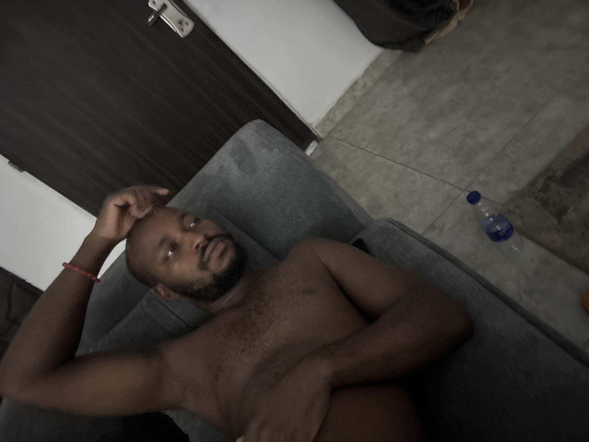 The image shows a man lying shirtless on a grey sofa in an indoor setting, likely in Iddo Tudunwada, Nigeria. The man has dark skin, a bald head, and a short, dark beard. He is looking directly at the camera with a relaxed or neutral expression. His right hand is placed near his head, while his left arm rests across his abdomen. A red beaded bracelet is visible on his left wrist.
The background reveals a simple domestic environment. To the left, a dark brown door with a silver handle and lock mechanism is visible. Adjacent to the door is a plain white wall. The floor is covered with large, light grey square tiles. A clear plastic water bottle with a blue cap rests on the tiled floor near the sofa. The overall lighting appears dim, suggesting the photo might have been taken in the evening, at night, or in a room with low artificial light. The man's posture indicates a moment of rest or relaxation. There is no visible text in the image.