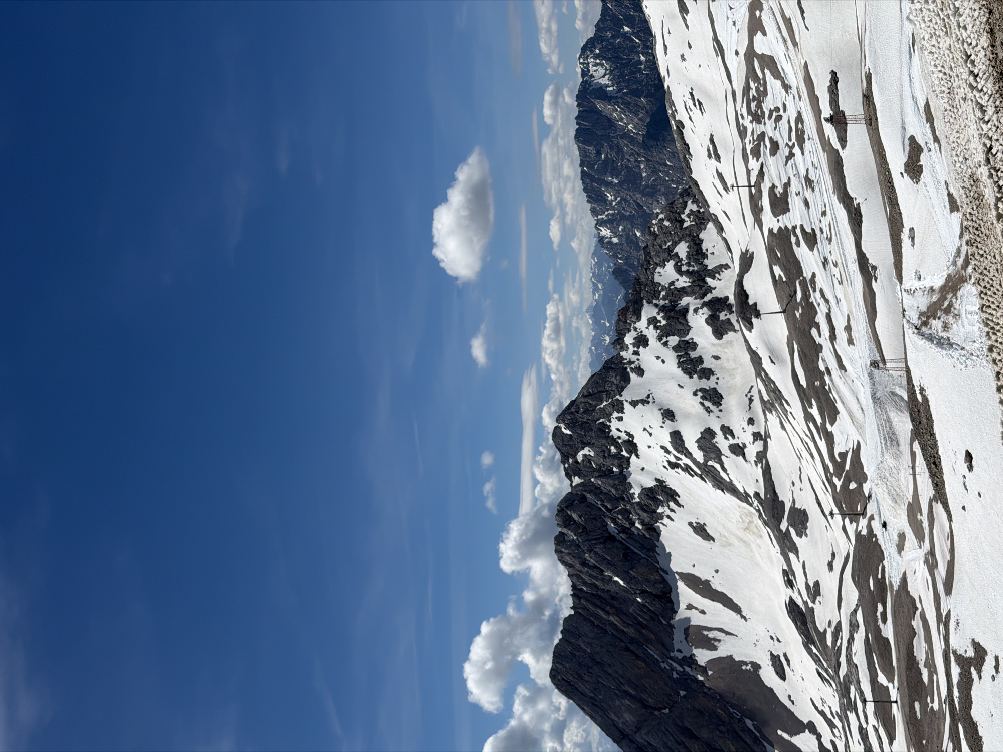 This image depicts a panoramic view of a snowy mountain landscape under a bright blue sky with scattered white clouds. The foreground is dominated by a rugged mountain range partially covered in snow, with exposed rocky sections and patches of bare earth. Several ski lift towers and cables are visible on the slopes, suggesting a ski resort. In the background, more mountain ranges are visible, some with snow-capped peaks. The overall scene conveys a sense of high altitude and winter or early spring conditions. The image appears to be taken from a high vantage point, possibly a mountain peak or a cable car, overlooking the expanse of the mountains. This landscape is characteristic of the Bavarian Alps, and specifically, the Zugspitze region near Garmisch-Partenkirchen, Germany, which is known for its prominent mountain scenery and ski resorts. There are no people or animals visible in the image.