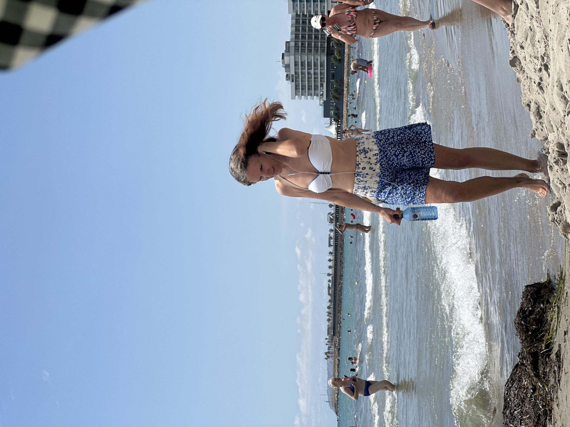 A medium shot shows a woman walking on a sandy beach in the foreground, her hair blowing in the wind. She is wearing a white bikini top and a blue and white floral skirt, and she is holding a plastic water bottle in her right hand. Her left hand is near her ear, and she appears to be wearing earbuds. The beach is populated with other people, some wading in the water and others sunbathing. In the background, there are buildings and palm trees, suggesting an urban coastal environment. The sky is clear and blue.