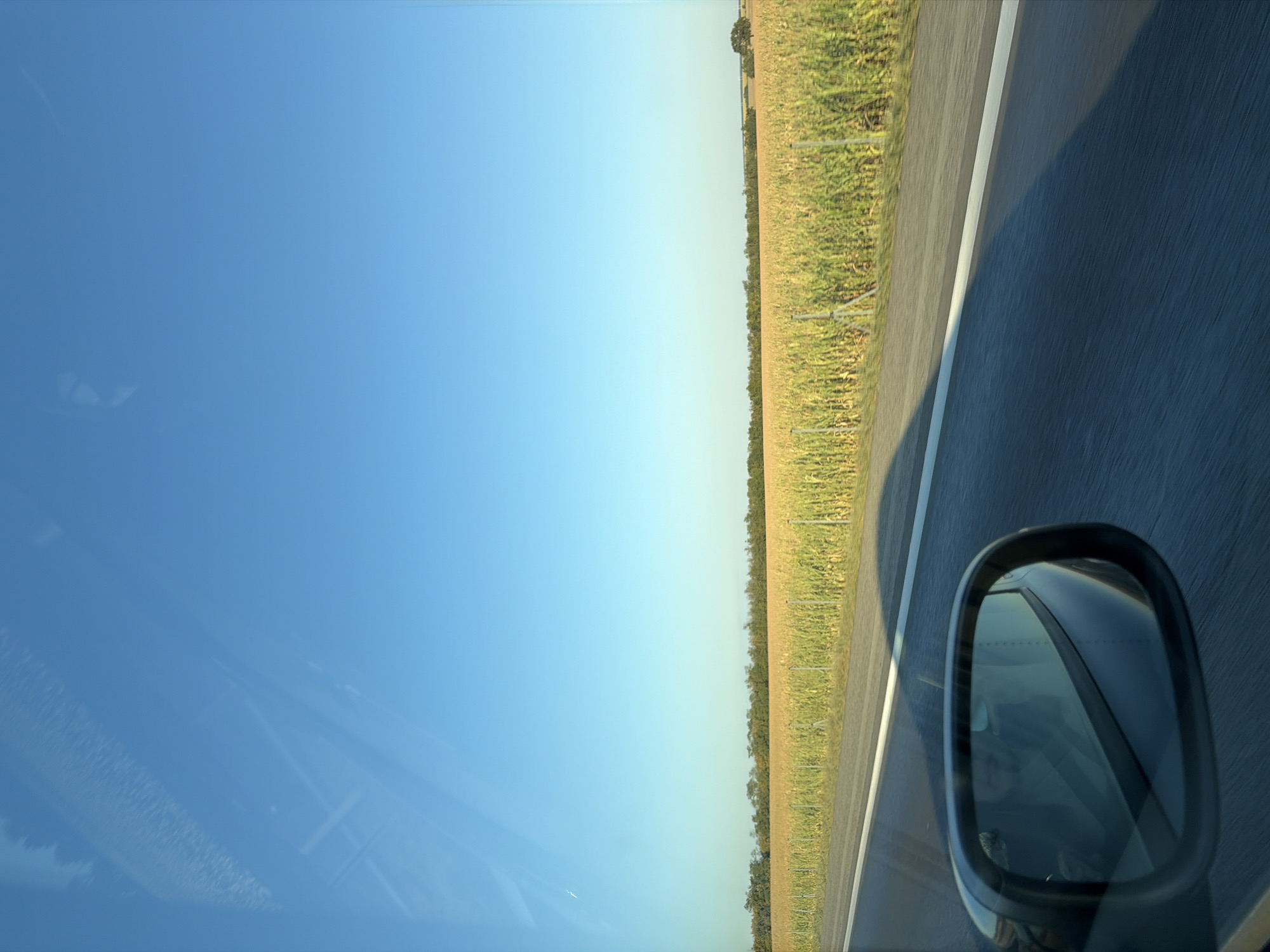 The image shows a view from inside a moving vehicle on a road. A bright blue sky dominates the upper portion of the frame. Below the sky, a vast field of tall, green and yellow corn stretches across the horizon, bordered by a line of trees. A white line marks the edge of the asphalt road in the foreground, which is blurred due to motion. A side-view mirror reflects the interior of the car, showing a person wearing a seatbelt. The scene suggests a rural setting, possibly in or near Pata, Slovakia, given the visual cues of agricultural land and a highway.