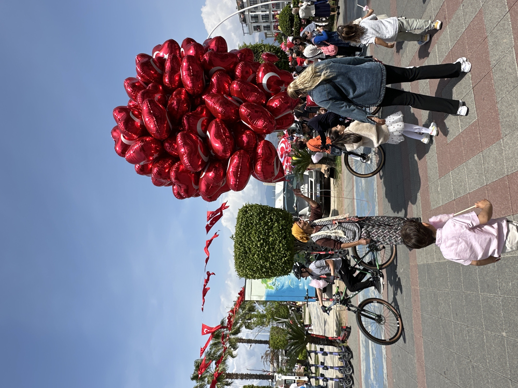 A wide-angle shot, rotated ninety degrees clockwise, presents a daytime scene outdoors in Fethiye, Turkey. A large cluster of red, heart-shaped balloons bearing the Turkish flag emblem (a white crescent moon and star) dominates the left side of the frame, contrasting against a clear, pale blue sky.
People are scattered across the scene, some walking, some riding bicycles. A woman with blonde hair, wearing a denim jacket and black pants, is visible in the middle-right of the image. Several children are nearby; one wears a white tutu and another is walking. A person wearing a yellow head covering is shown riding a bicycle near a green shrub. Another person in a pink shirt is visible.
Along the street are trees decorated with Turkish flags, providing a sense of location. Parked electric scooters line the curb. Buildings can be seen in the background. The ground is paved with red and grey bricks.