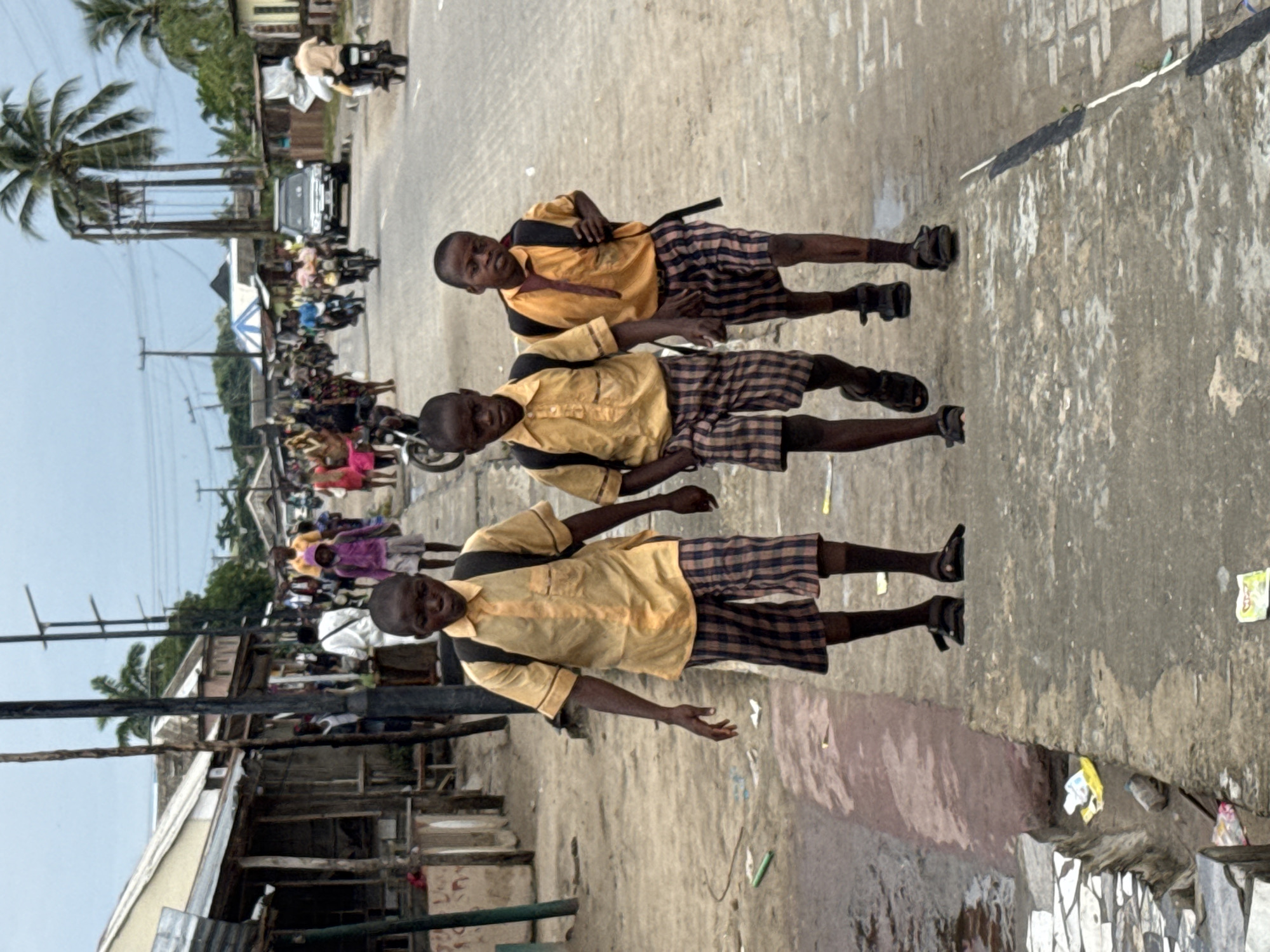 Three schoolboys in yellow shirts and plaid shorts walk along a busy street in Badagry, Nigeria. They wear backpacks and sandals. The street is paved but uneven, with visible trash and debris. In the background, a crowd of people and vehicles, including motorcycles and a car, move along the road. Palm trees and power lines are visible in the distance. Buildings line one side of the street, with corrugated metal and wood structures. The sky is overcast.