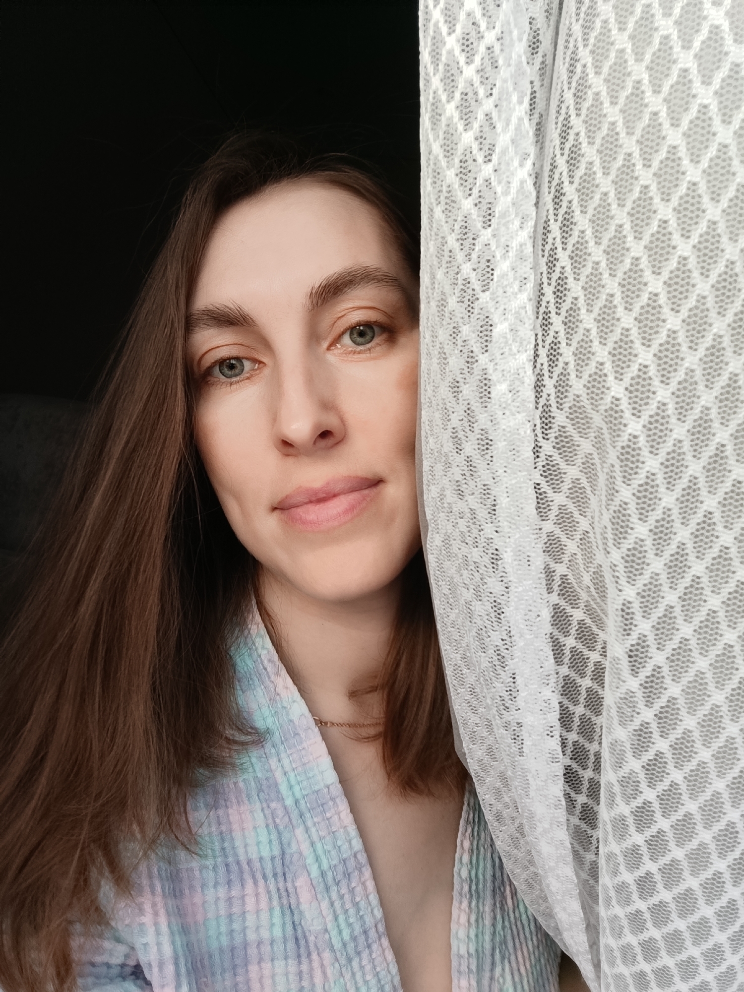 A close-up, chest-level portrait shot captures a female subject indoors in what is specified as Chaikovsky, Russia, though no distinguishing geographical features are discernible in the image. The subject is a light-skinned individual with long, straight brown hair parted on the viewer's left, flowing over her shoulder. Her face is directed towards the camera, featuring light blue or green eyes, natural eyebrows, and a slight, closed-lip smile. A small blemish or mark is visible on her right cheek. She is wearing a pastel-colored (light blue, pink, purple, white) plaid-patterned garment, possibly a bathrobe, made of a textured, waffle-knit fabric, with a thin gold-colored chain necklace visible around her neck. The right side of her body, from the viewer's perspective, is partially obscured by a translucent white curtain with a distinct hexagonal or honeycomb mesh pattern. The background on the left is dark and indistinct, while the curtain suggests proximity to a window, providing soft, diffused lighting on the subject's face.