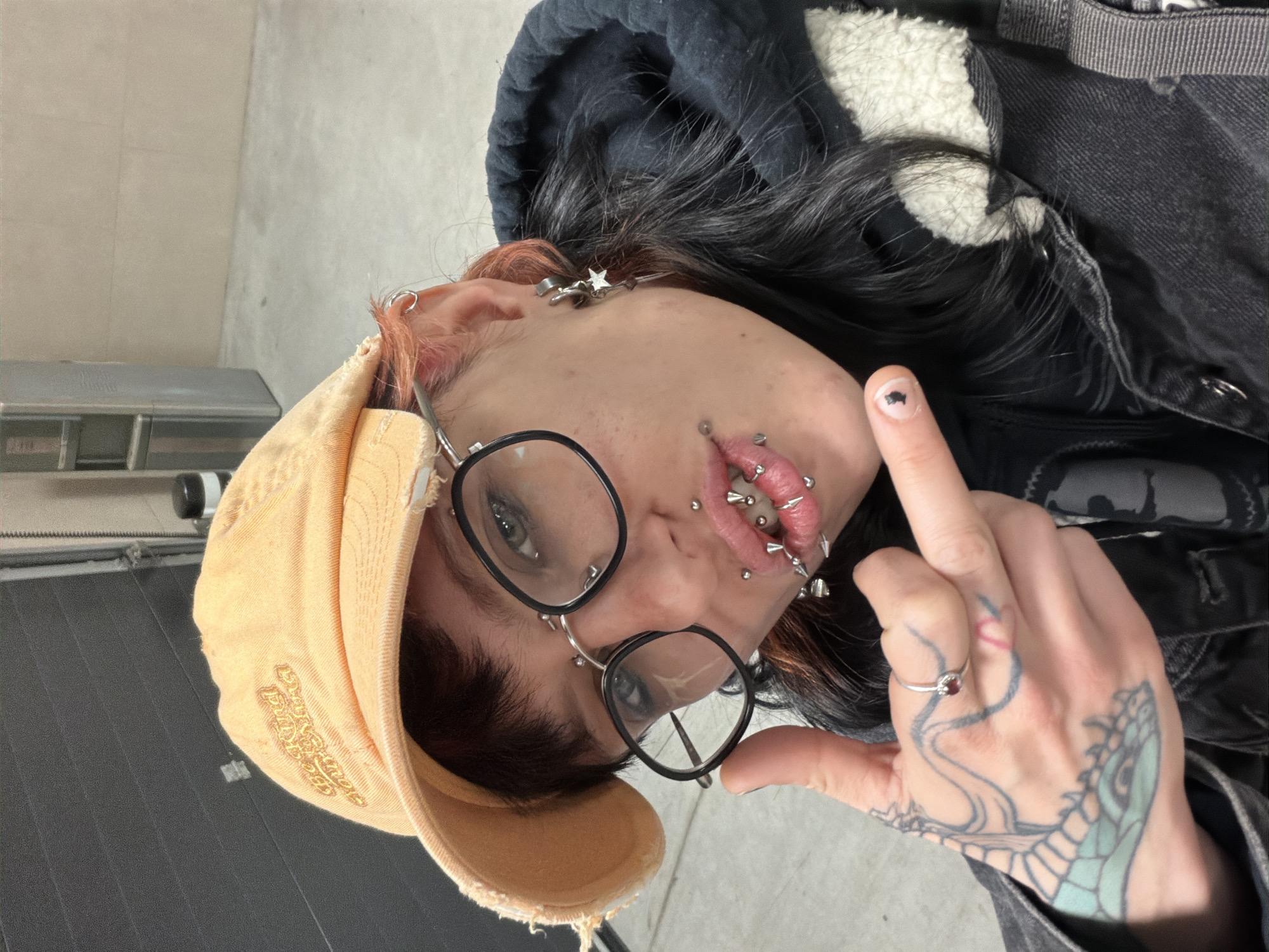 A person is indoors, wearing a yellow baseball cap, round black glasses, a dark hoodie, and a black denim jacket with a white fleece lining. They have multiple lip piercings and facial piercings, including studs and rings, and several ear piercings. The person is displaying their middle finger. Their hand is tattooed, and they are wearing a ring on their ring finger. The background shows a light-colored wall and a section of what appears to be a garage door. There is no indication of location in Busmantsi, Bulgaria.