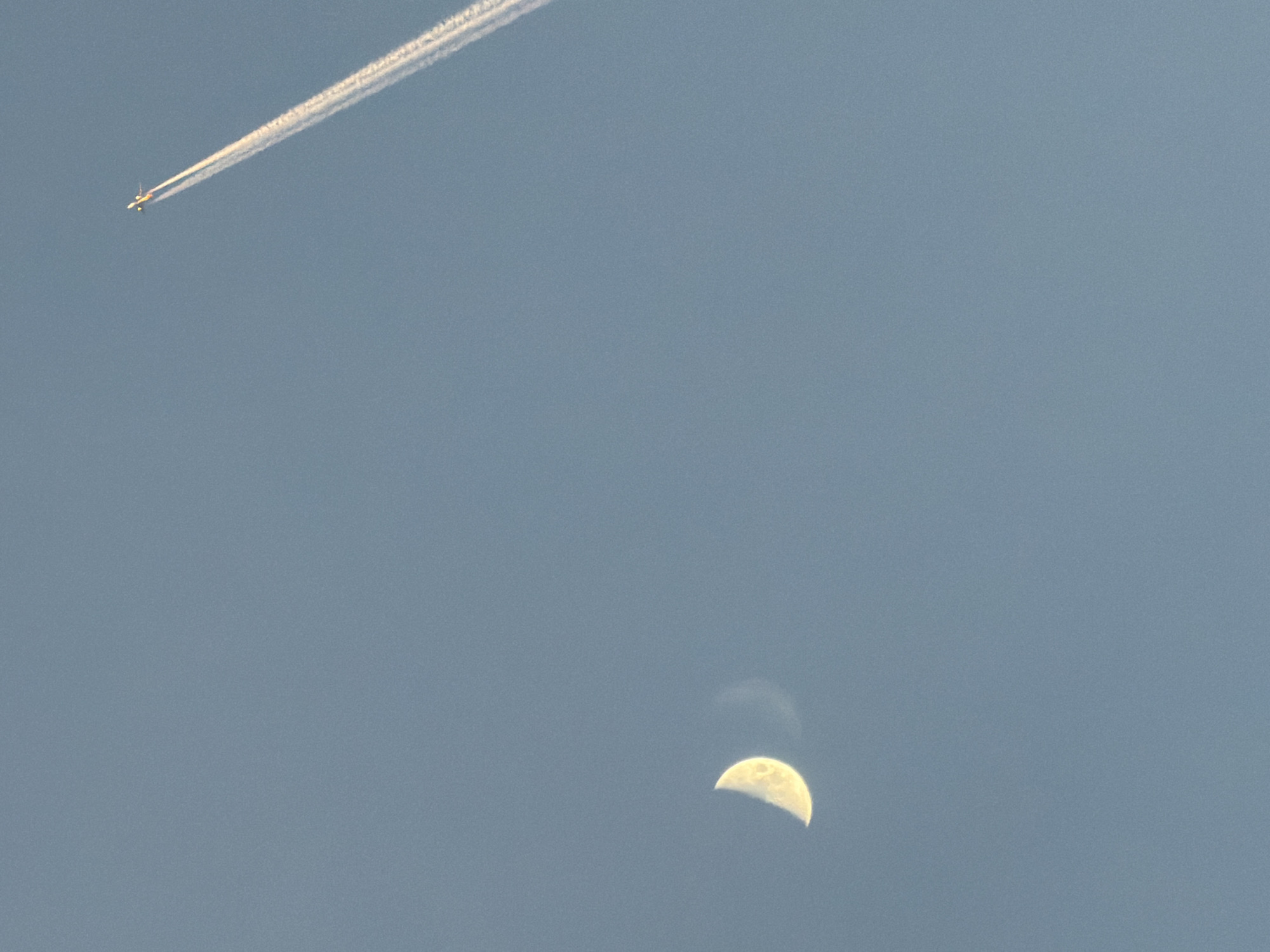 The image depicts a daytime sky in Кадряково, Russia. In the upper left quadrant, a commercial jet aircraft is leaving a white contrail. In the lower right quadrant, a crescent moon is visible.