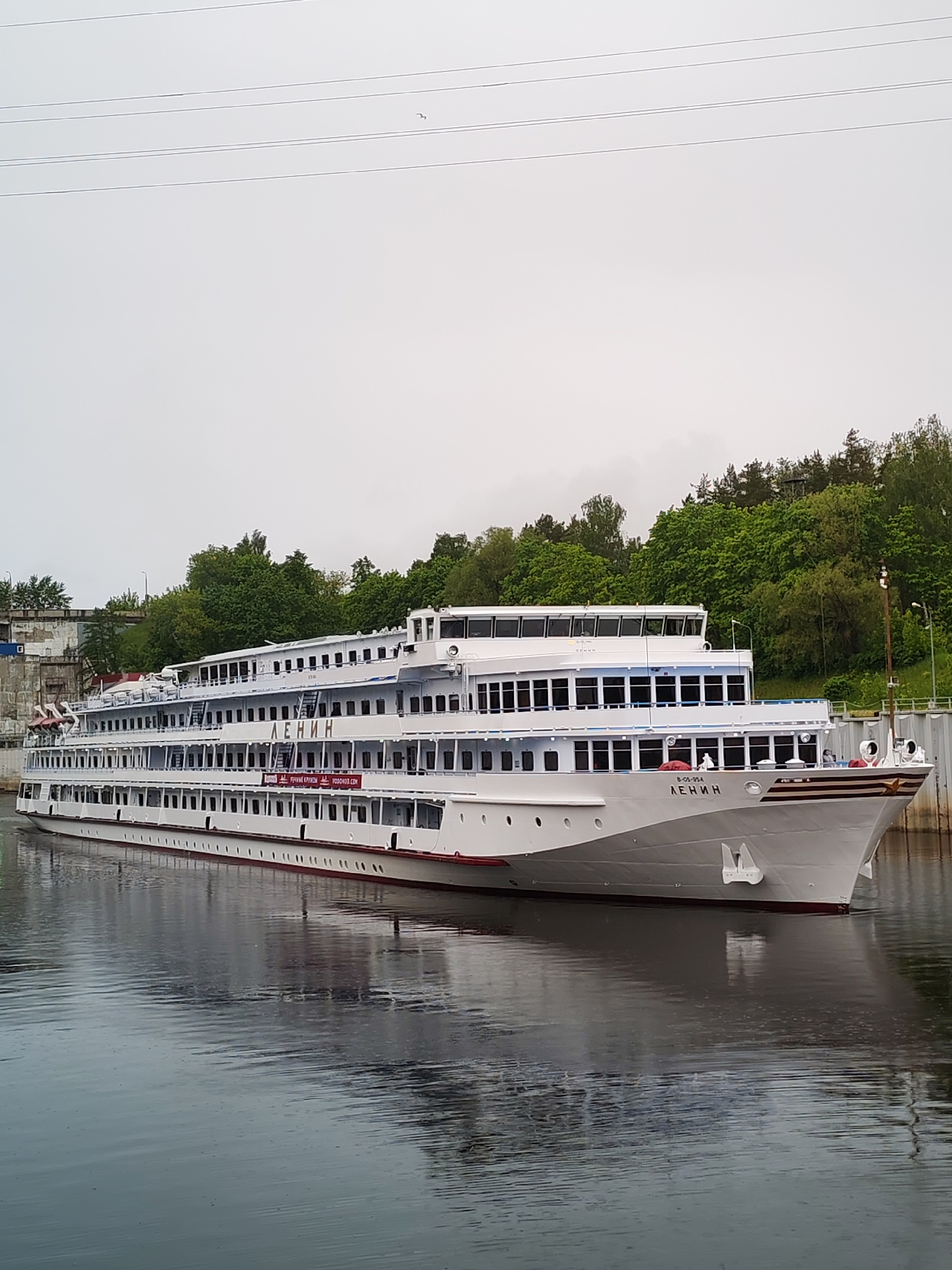 A large white passenger ship with multiple decks is docked along a riverbank. The ship has the word "ЛЕНИН" (Lenin) prominently displayed on its side. Green trees and foliage line the riverbank behind the ship. A gray, overcast sky is visible above. The water is calm and reflects the ship and surrounding environment. The location is in the city of Bol'shie Mkhi, Russia.