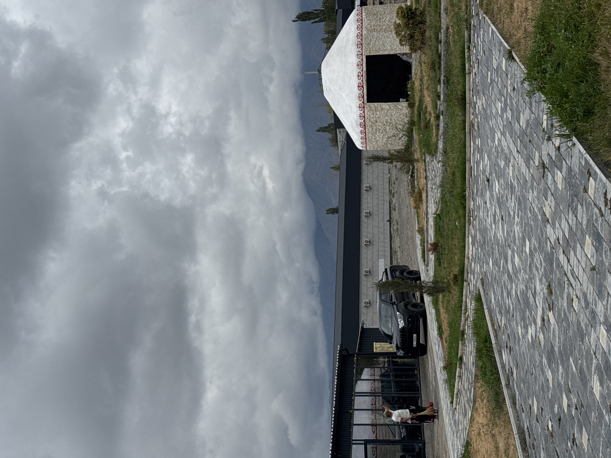 In Balykchy, Kyrgyzstan, under an overcast sky, a scene unfolds featuring a paved walkway leading towards modern buildings and a traditional yurt. A person in a white t-shirt and a cap is visible on the left, holding a broom and standing near a dark-colored pickup truck. The truck is parked in front of a building with large glass windows. To the right, a stone yurt with a white roof and red decorative trim stands open, revealing a dark interior. In the background, a long, low-rise building with air conditioning units is visible, and beyond that, mountains are shrouded in mist and clouds. The ground is a mix of dry grass and the grey patterned paving stones.
