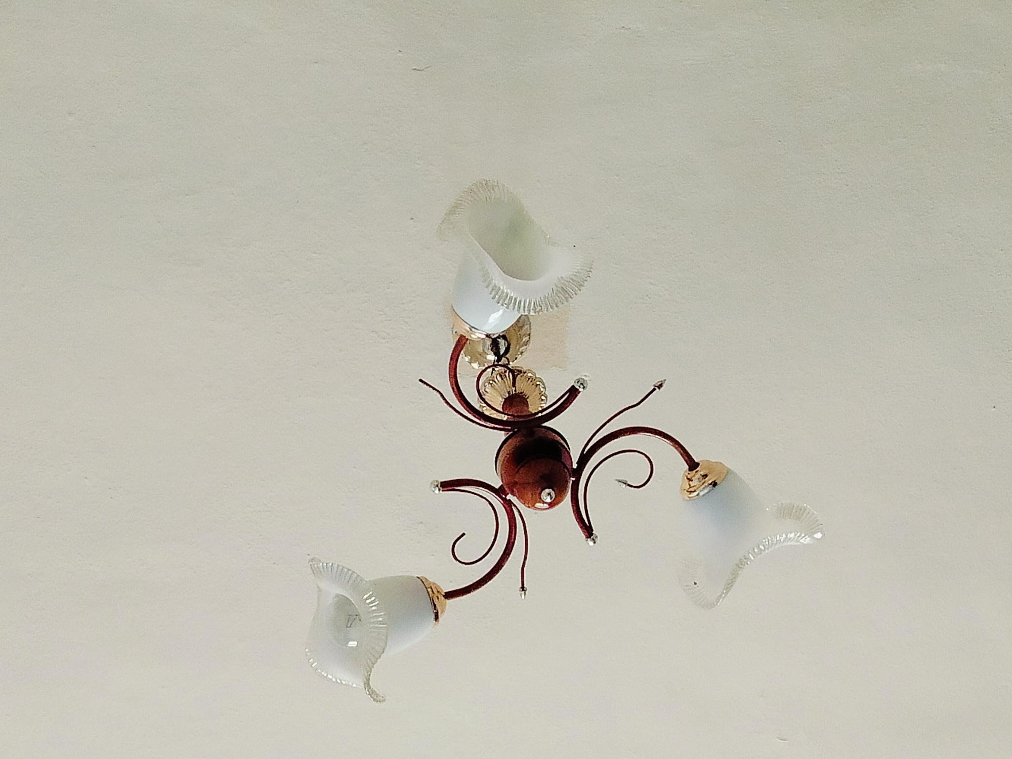 An ornate ceiling light fixture is pictured against a plain white ceiling. The fixture has three arms, each holding a white, bell-shaped glass shade with a ruffled edge. The arms are made of dark red, curved metal, with some decorative gold accents. A central, dark red, spherical element connects the arms. The image does not contain any contextual information that would allow for the identification of its location in the city of Көлкент, Kazakhstan.