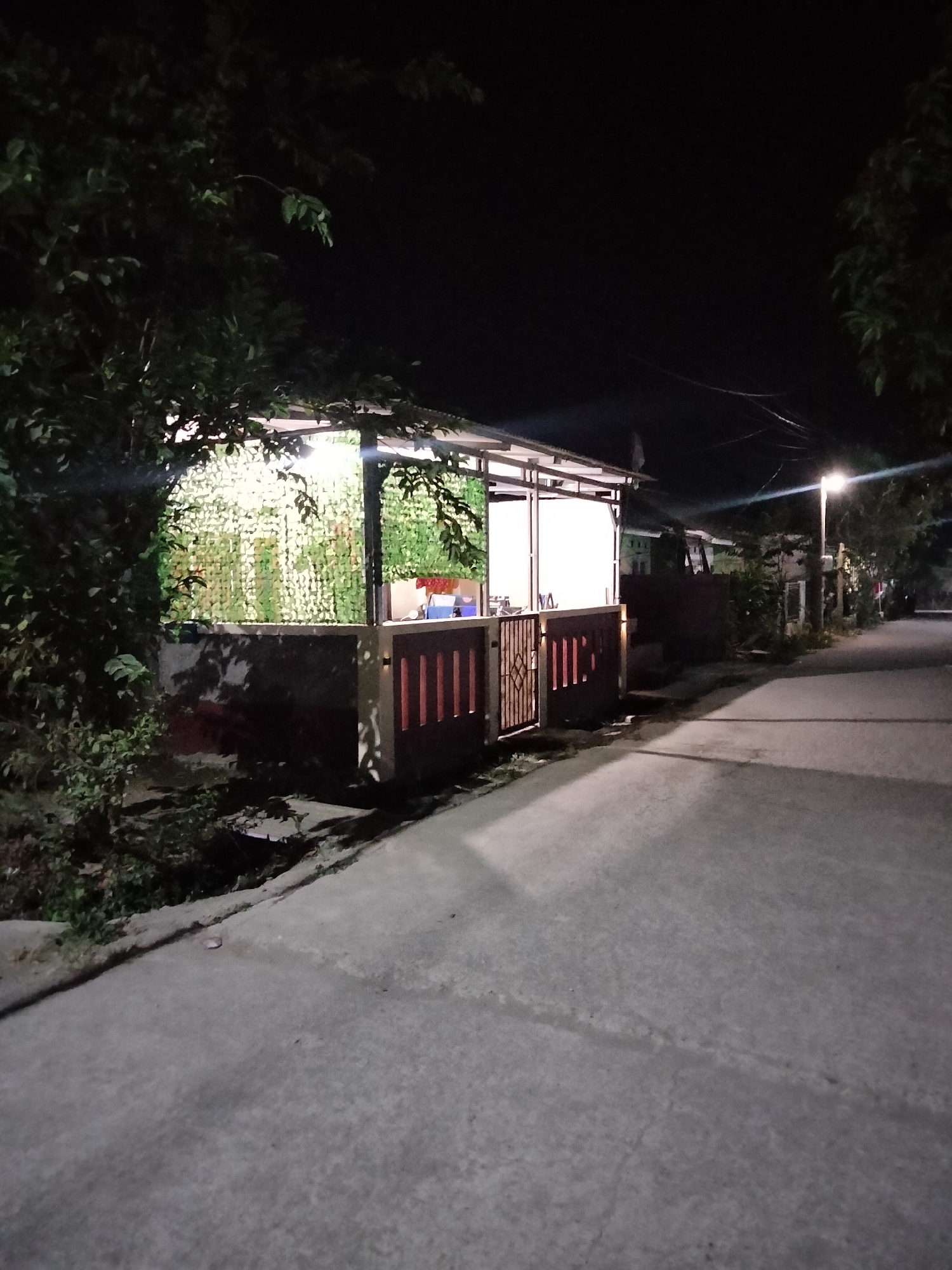 The image depicts a night scene in Cariu, Indonesia. In the foreground, a paved road curves towards the right, illuminated by a streetlamp on the right side. Adjacent to the road, on the left, is a structure with an open-air shelter. The shelter has a roof supported by poles and is partially enclosed by a dark-colored fence or wall with vertical slats. Behind the fence, a green, net-like material hangs, with some lighting visible behind it, suggesting the interior of the shelter is illuminated. Trees with dark foliage frame the left and right sides of the image. The overall environment appears to be a residential or commercial area at night, with artificial lighting providing visibility.